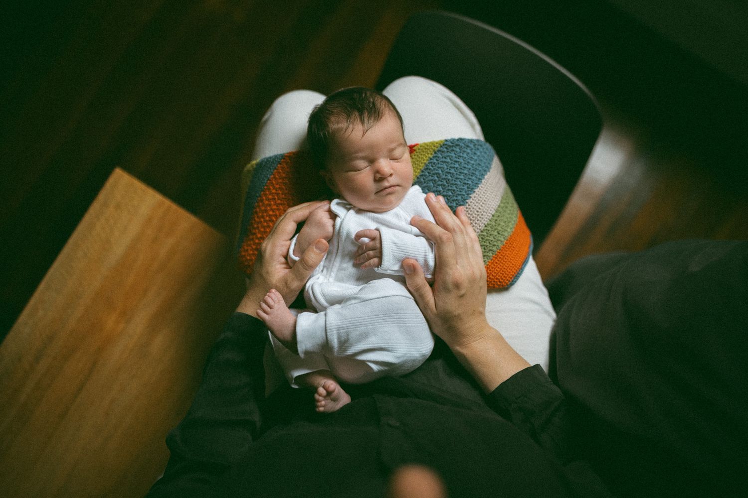Newborn baby sleeping peacefully in a white chair while a parent's hands gently support them.
