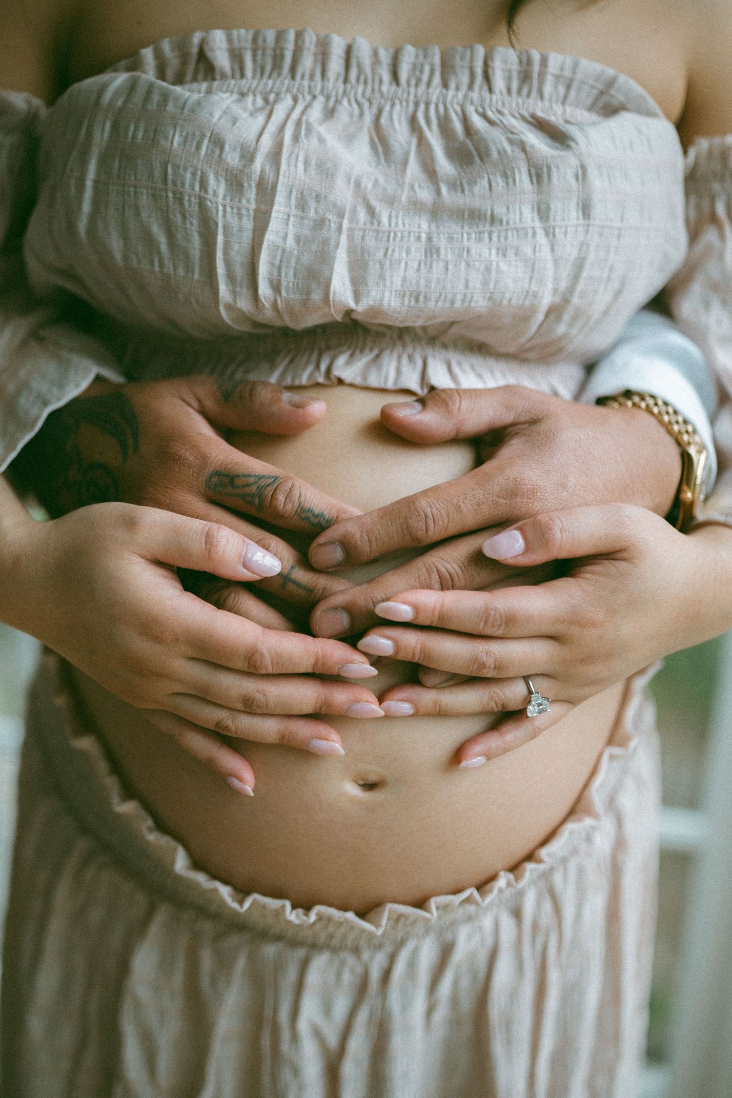 Pregnant woman in off-shoulder top with two pairs of hands cradling baby bump, engagement ring visible.