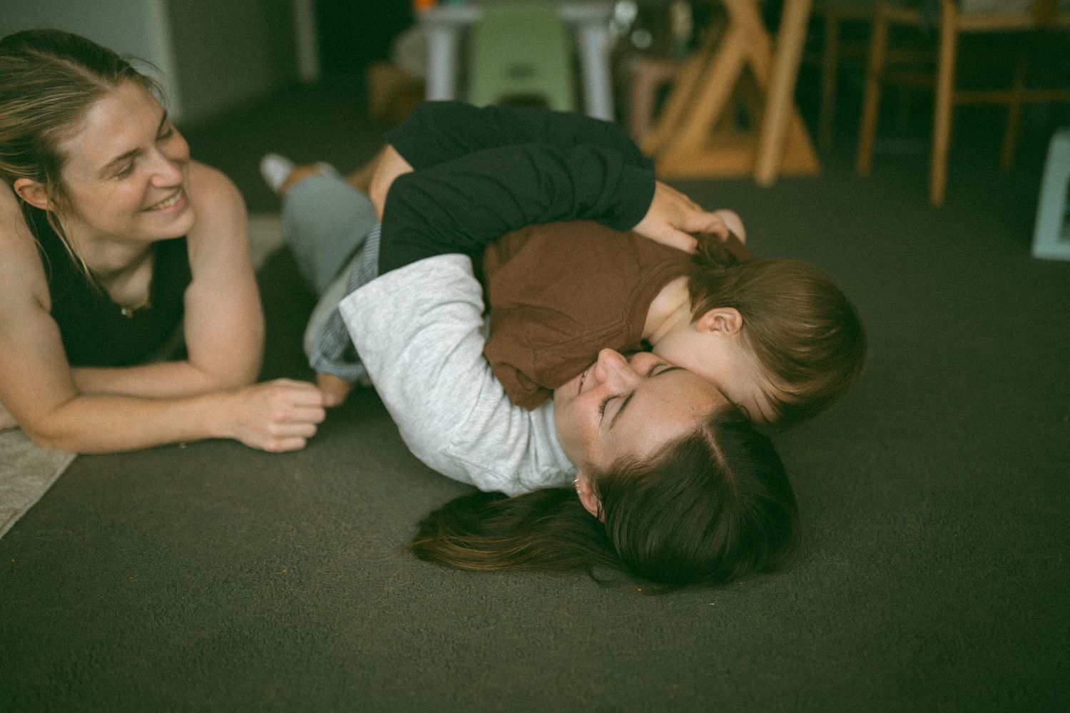 Two women playing and laughing on the floor, one lying down with a child on her chest in a cozy indoor setting.