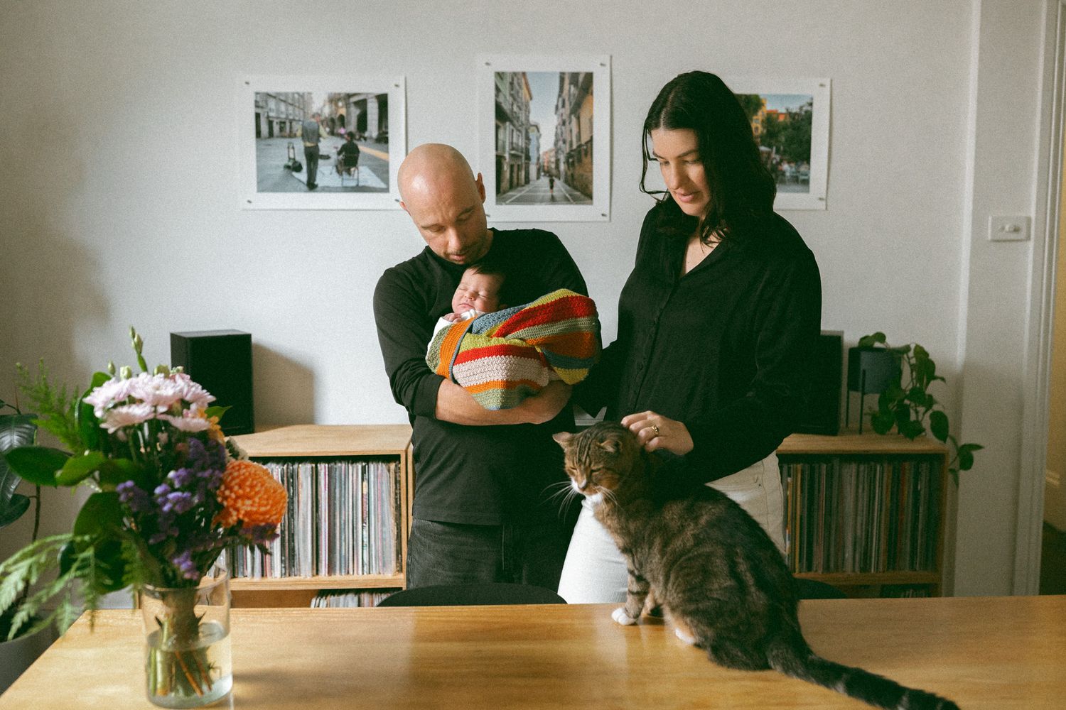 A couple with a newborn baby and a tabby cat on a wooden table, standing in a cozy apartment with vinyl records and photos.