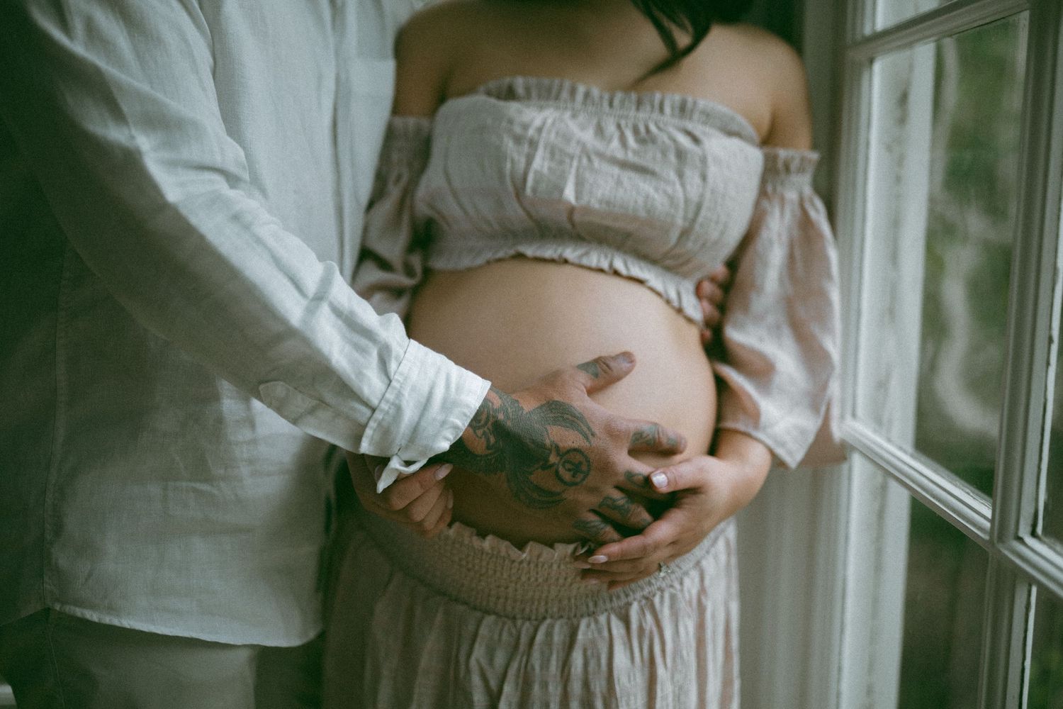 Pregnant woman in off-shoulder dress with tattoos, cradling baby bump beside partner near window.