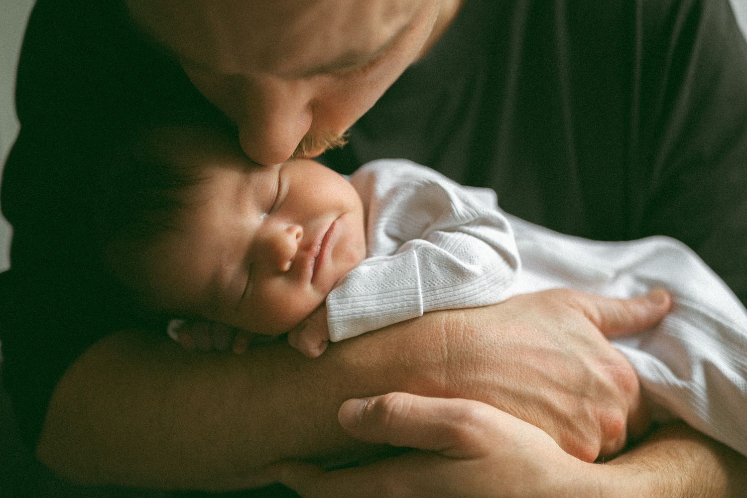 Father gently kissing his sleeping newborn baby, cradling the infant tenderly in his hands.