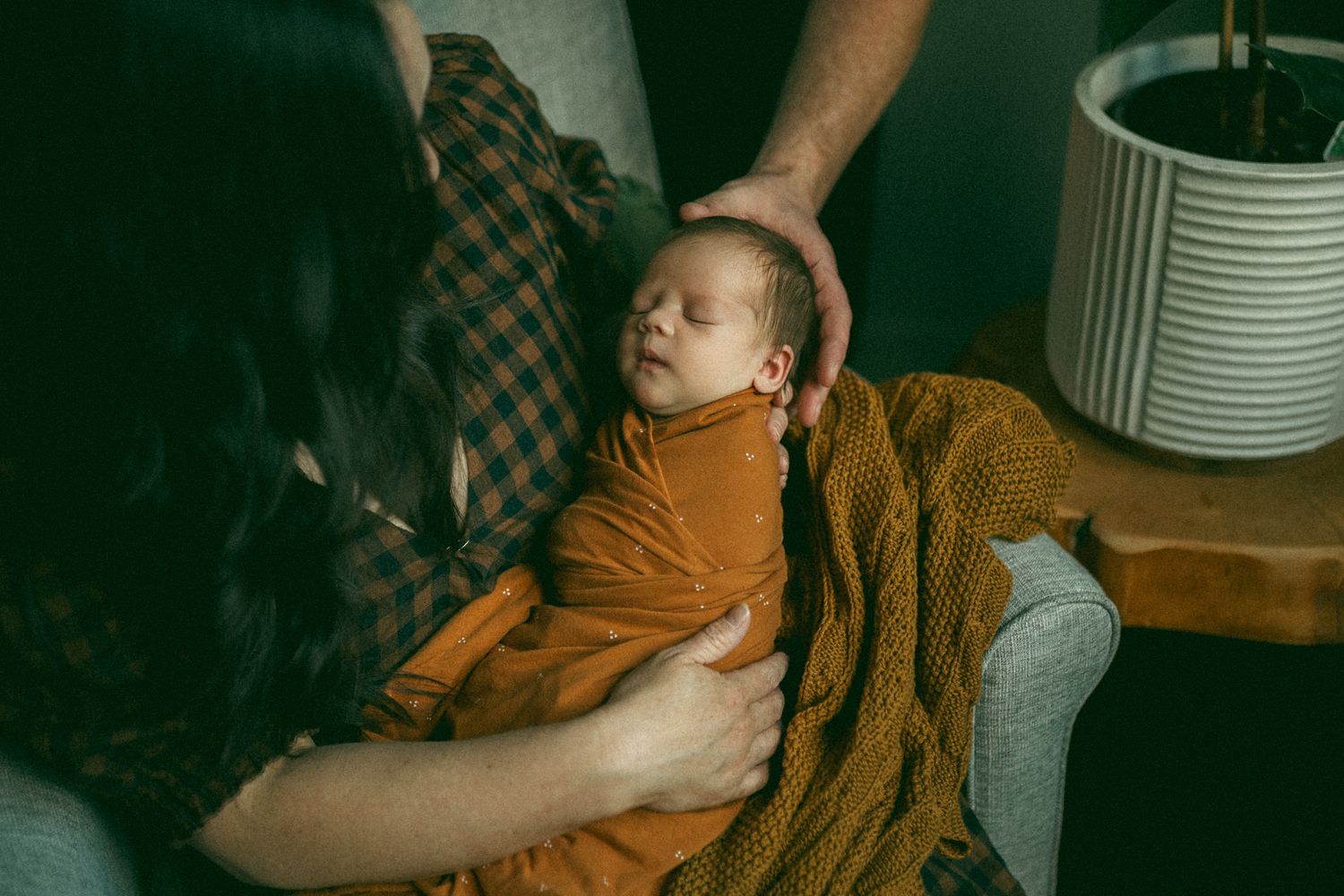 Newborn baby sleeping peacefully wrapped in mustard yellow blanket while parents hold and cradle infant in cozy home setting.
