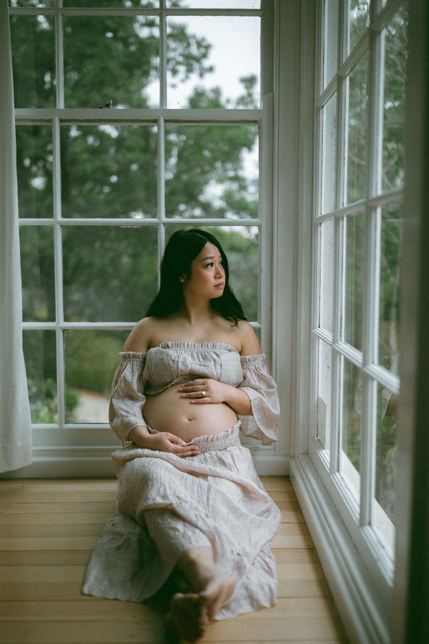 Pregnant woman in off-shoulder floral dress sits by large window, cradling baby bump, gazing outside thoughtfully.