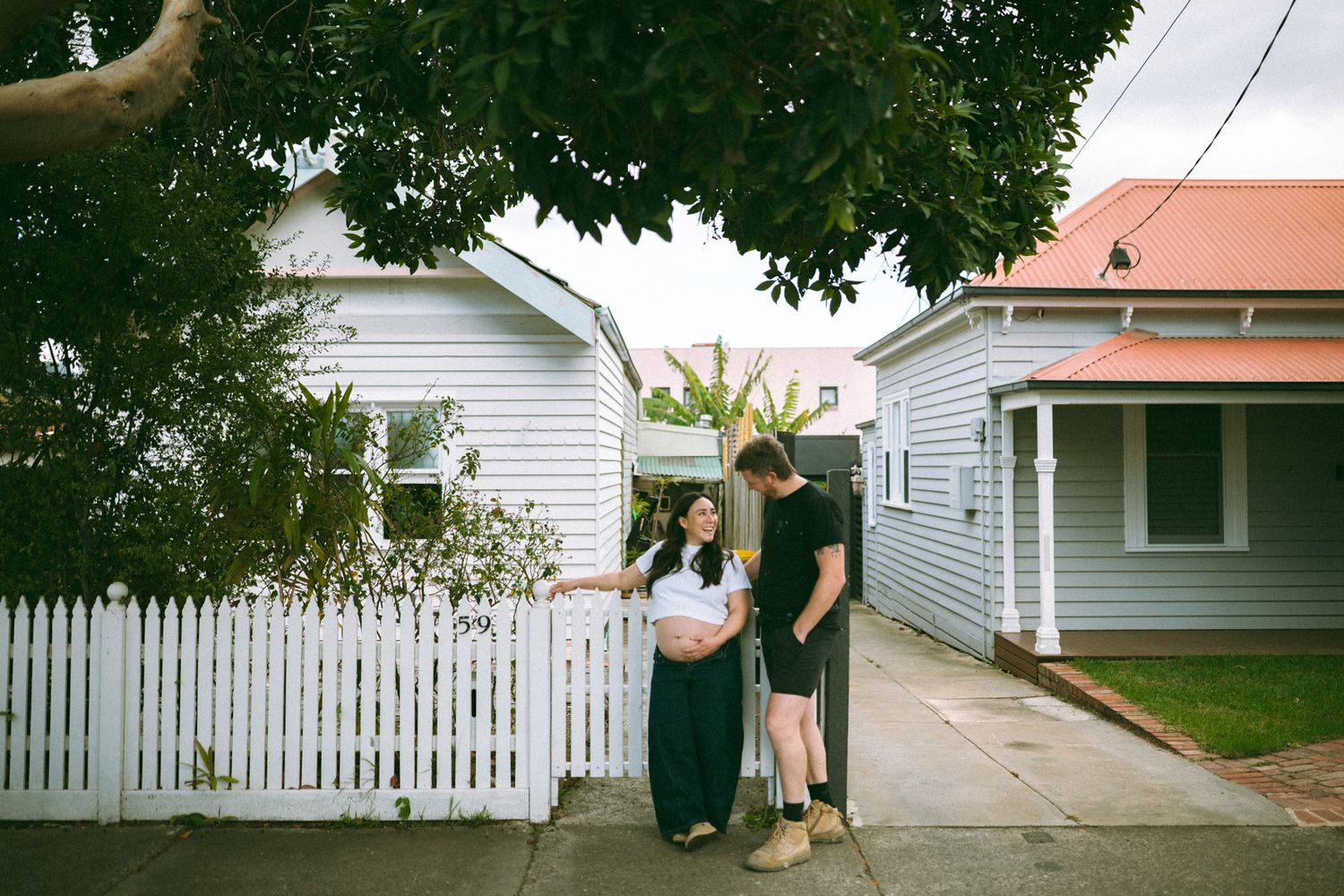 Pregnant couple standing on sidewalk in front of white picket fence and charming weatherboard homes on a cloudy day.