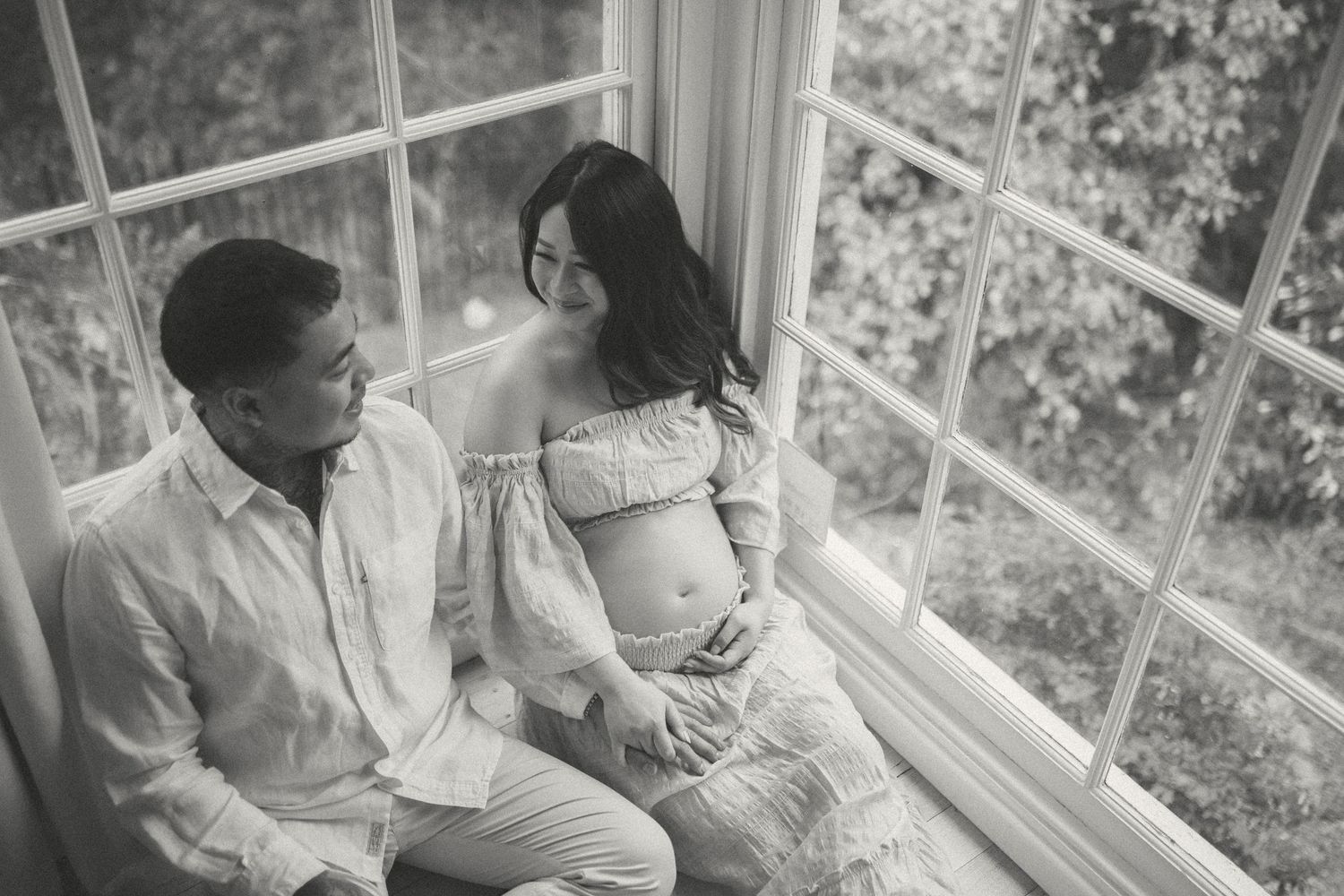 Black and white maternity photo of a couple sitting by a large window, smiling at each other, woman cradling her baby bump.