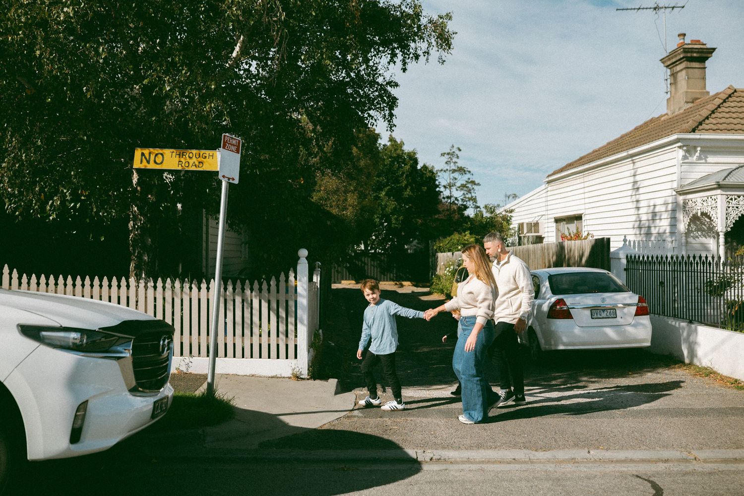 Family of three holding hands near parked cars on a suburban street beside a white picket fence on a sunny day.