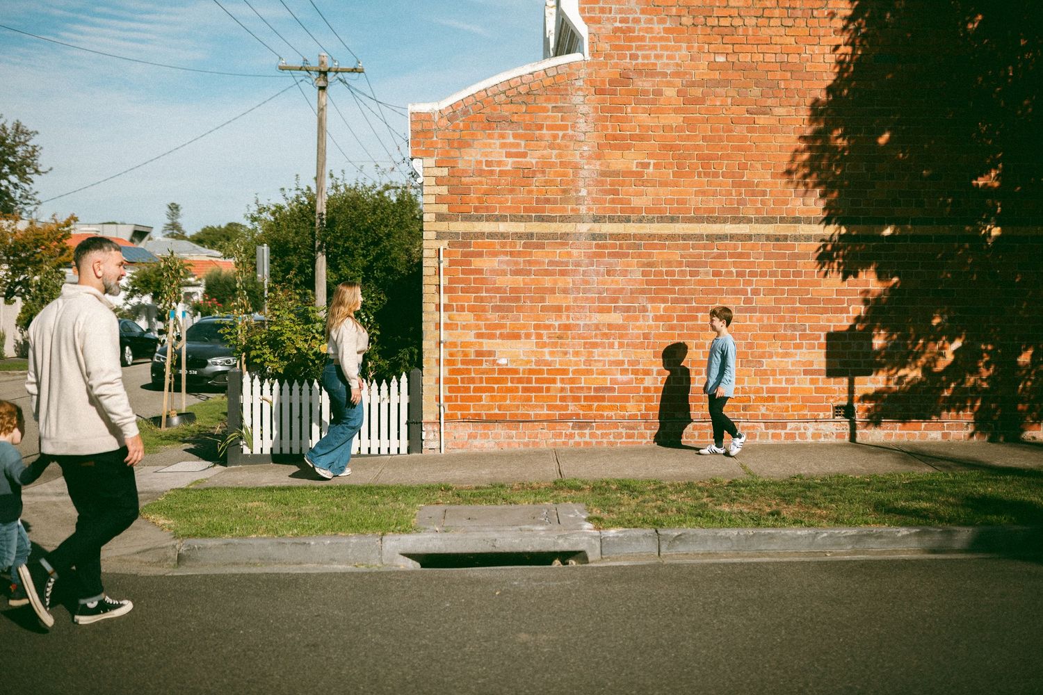 People walking on a suburban street beside a red brick building on a sunny day.