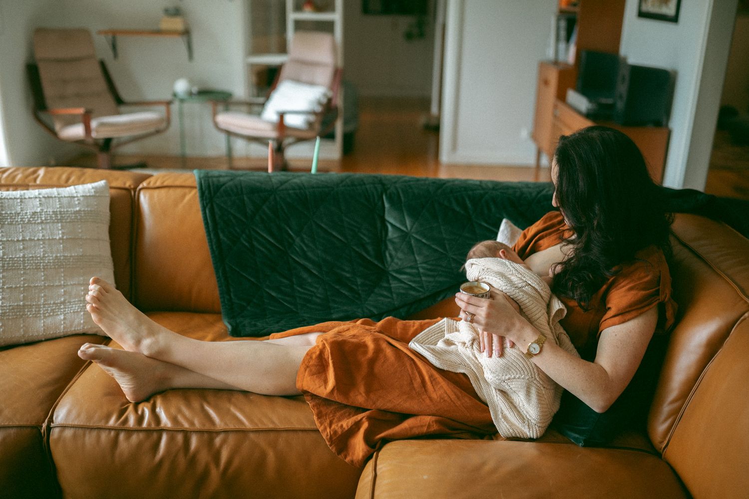 A woman in an orange dress reclines on a leather sofa, cradling a newborn baby in a cozy living room.