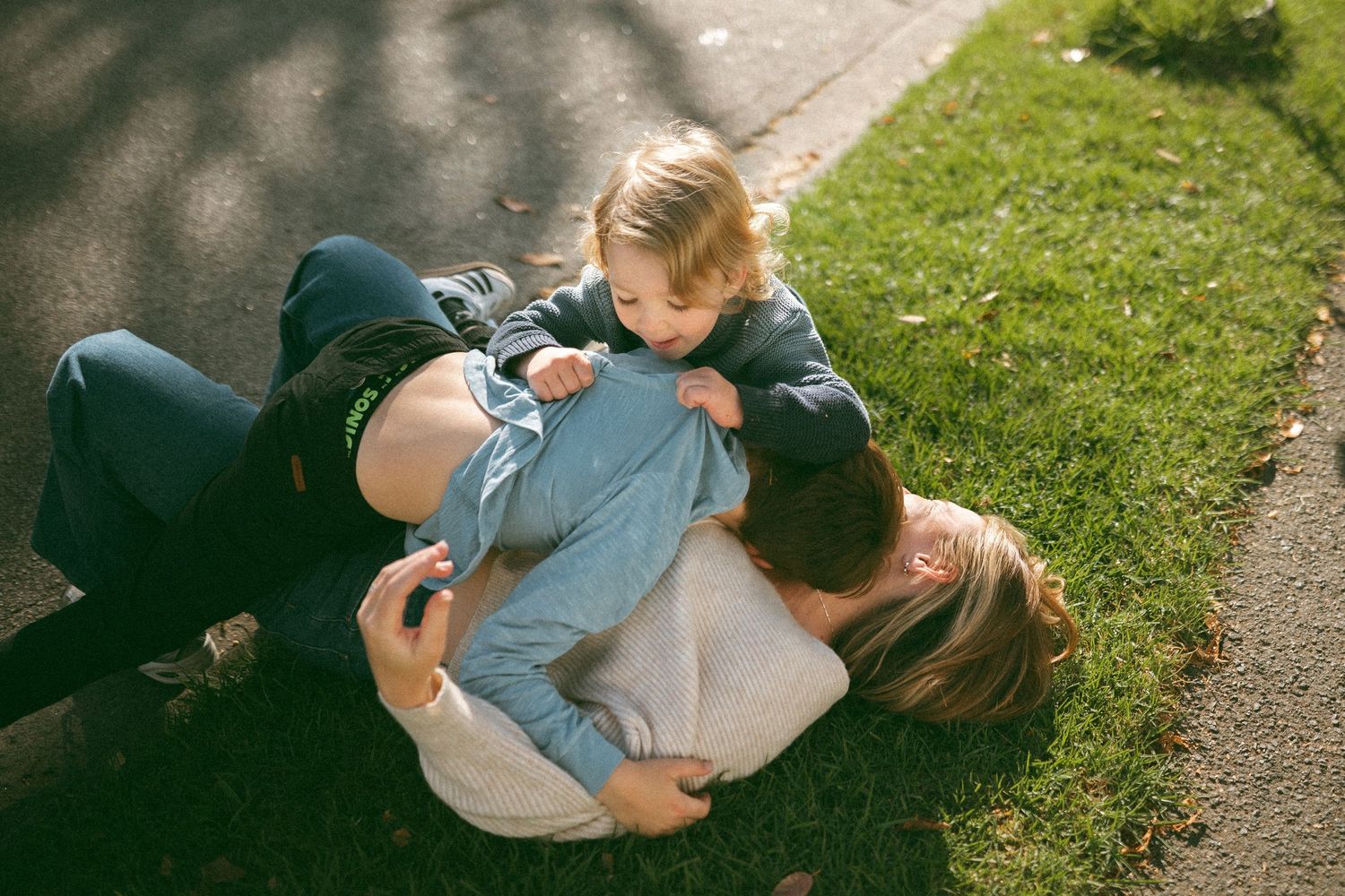 Two adults and a toddler playfully wrestling on a sunny lawn beside a paved path.