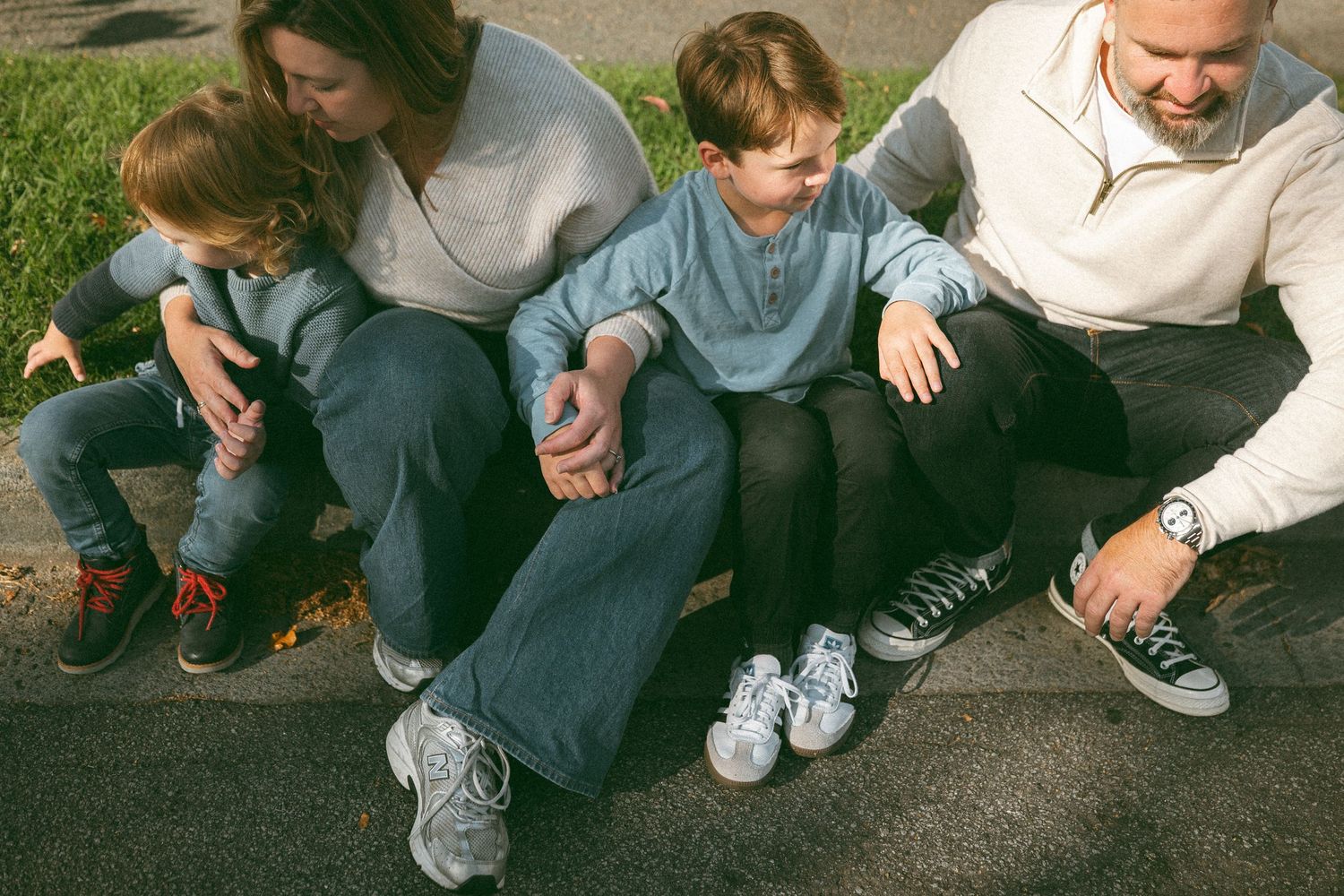 Family of four sitting together outdoors on a curb, two young boys with parents, wearing casual clothes and sneakers.