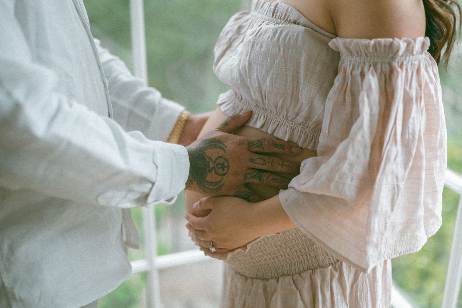 Couple tenderly cradling a pregnant belly, man with tattoed hand in white shirt, woman in off-shoulder dress.