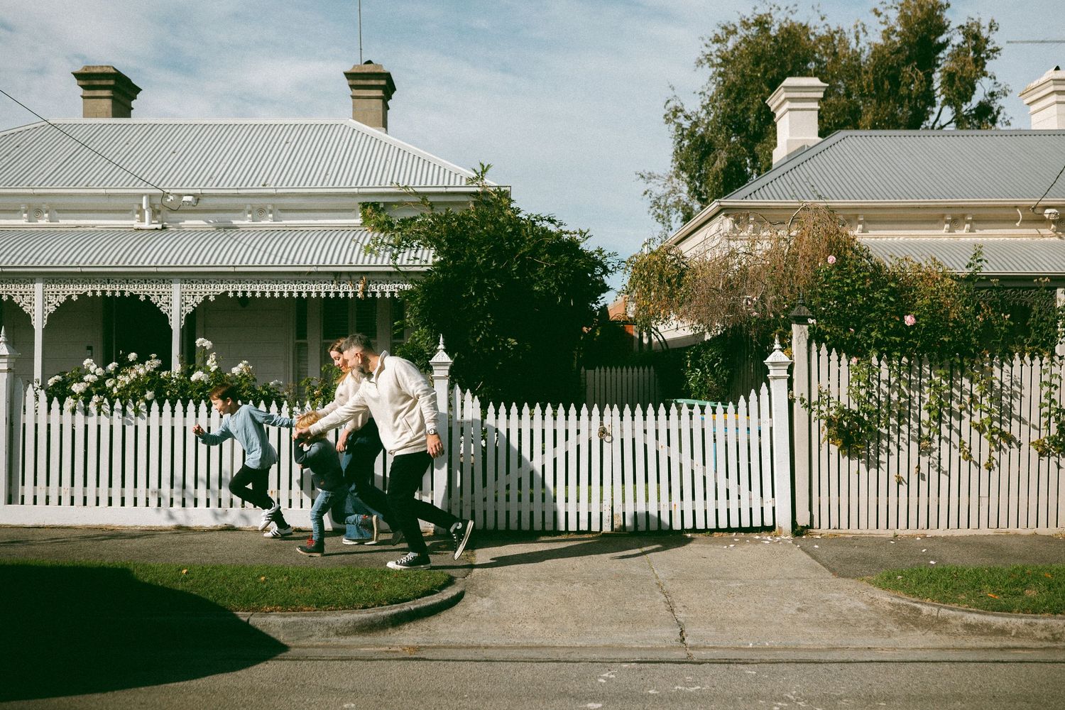 People riding a tandem bike past a white picket fence and charming Victorian homes on a sunny suburban street.