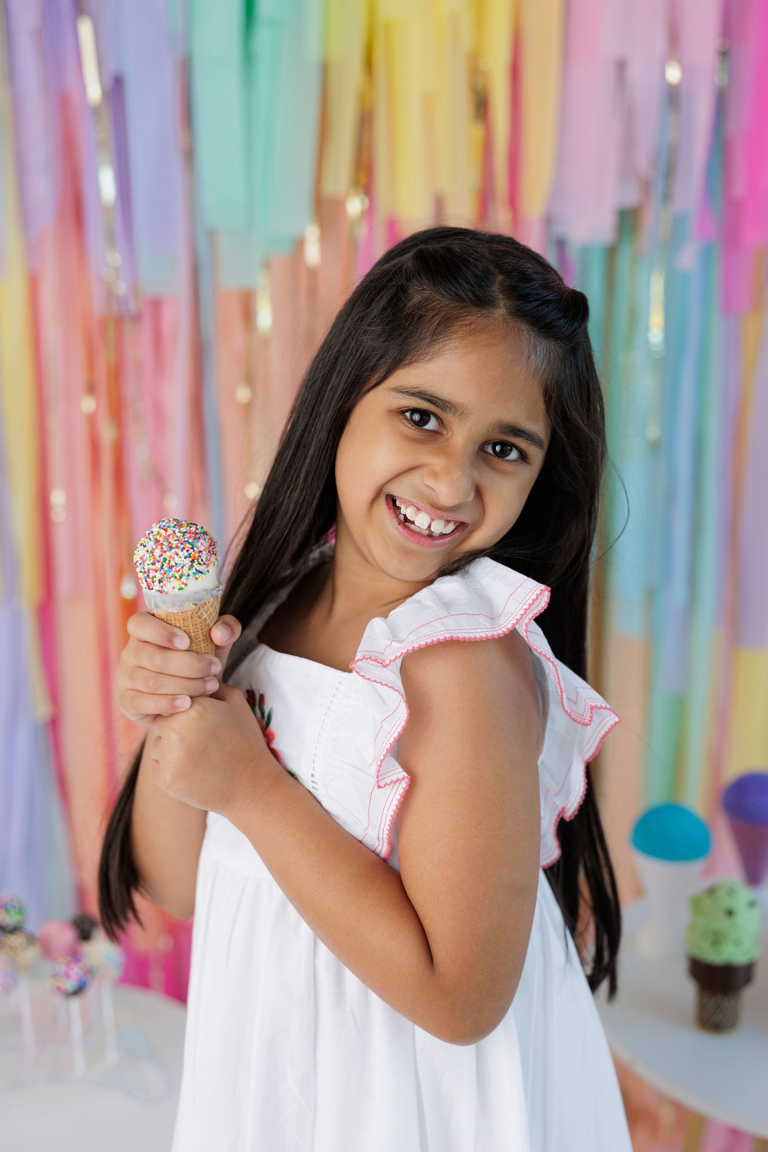 Young girl in white dress holding a cake pop against colorful streamer background at a party celebration.