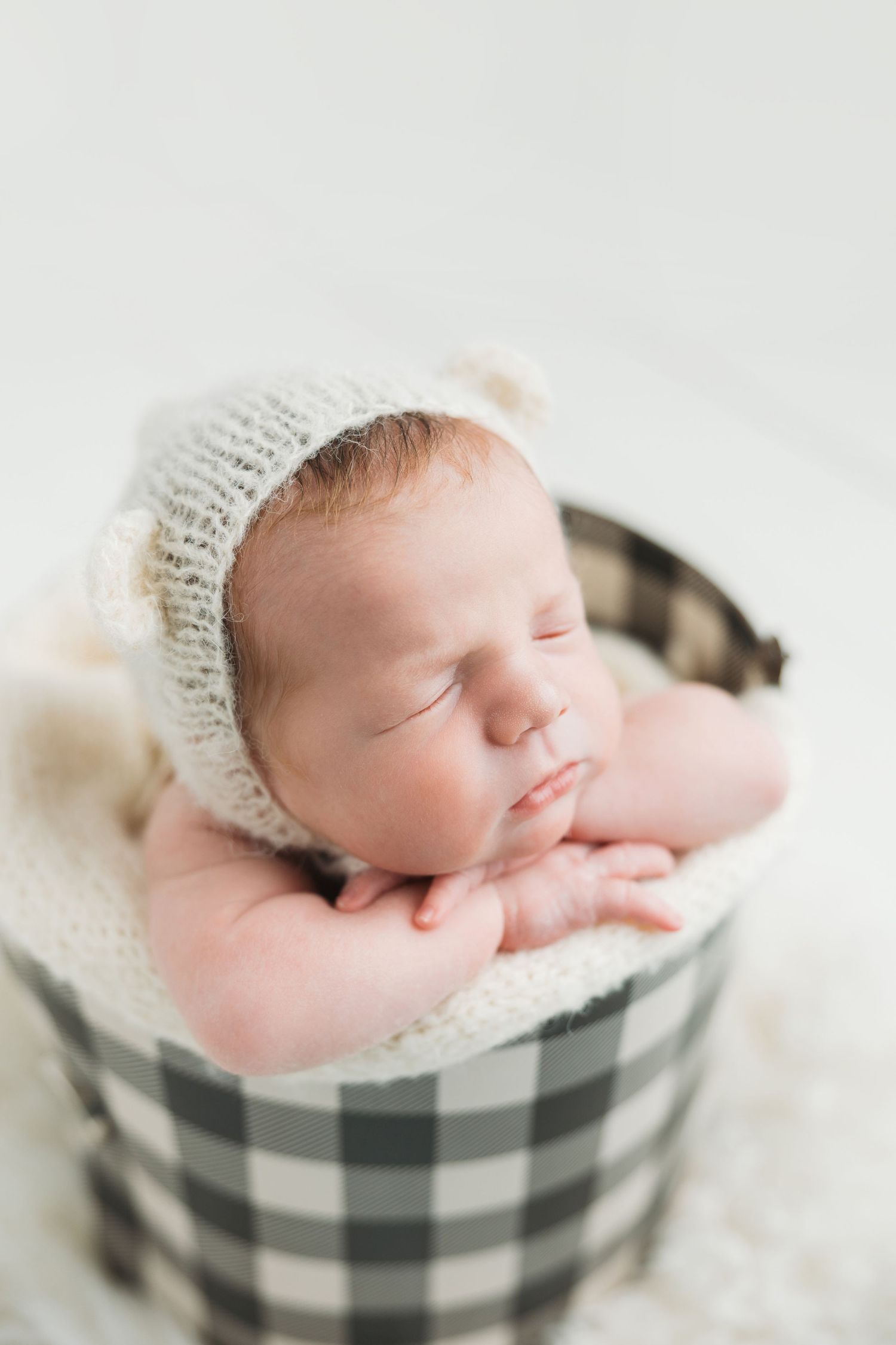 Sleeping newborn wearing knit bonnet rests peacefully in black and white checkered basket with soft bedding.
