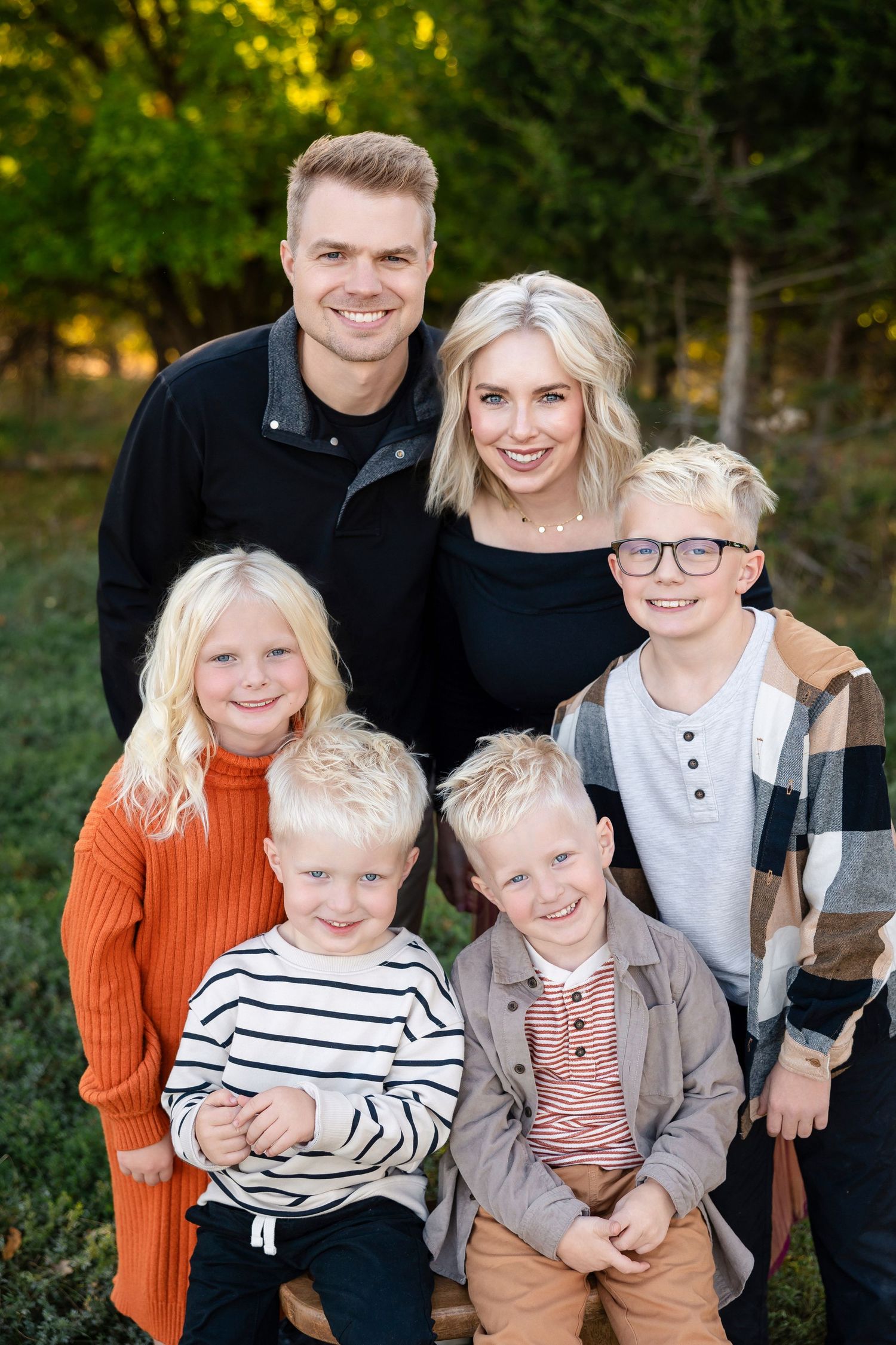 Outdoor family portrait of five people wearing casual fall clothing posed together in front of trees with golden sunlight.