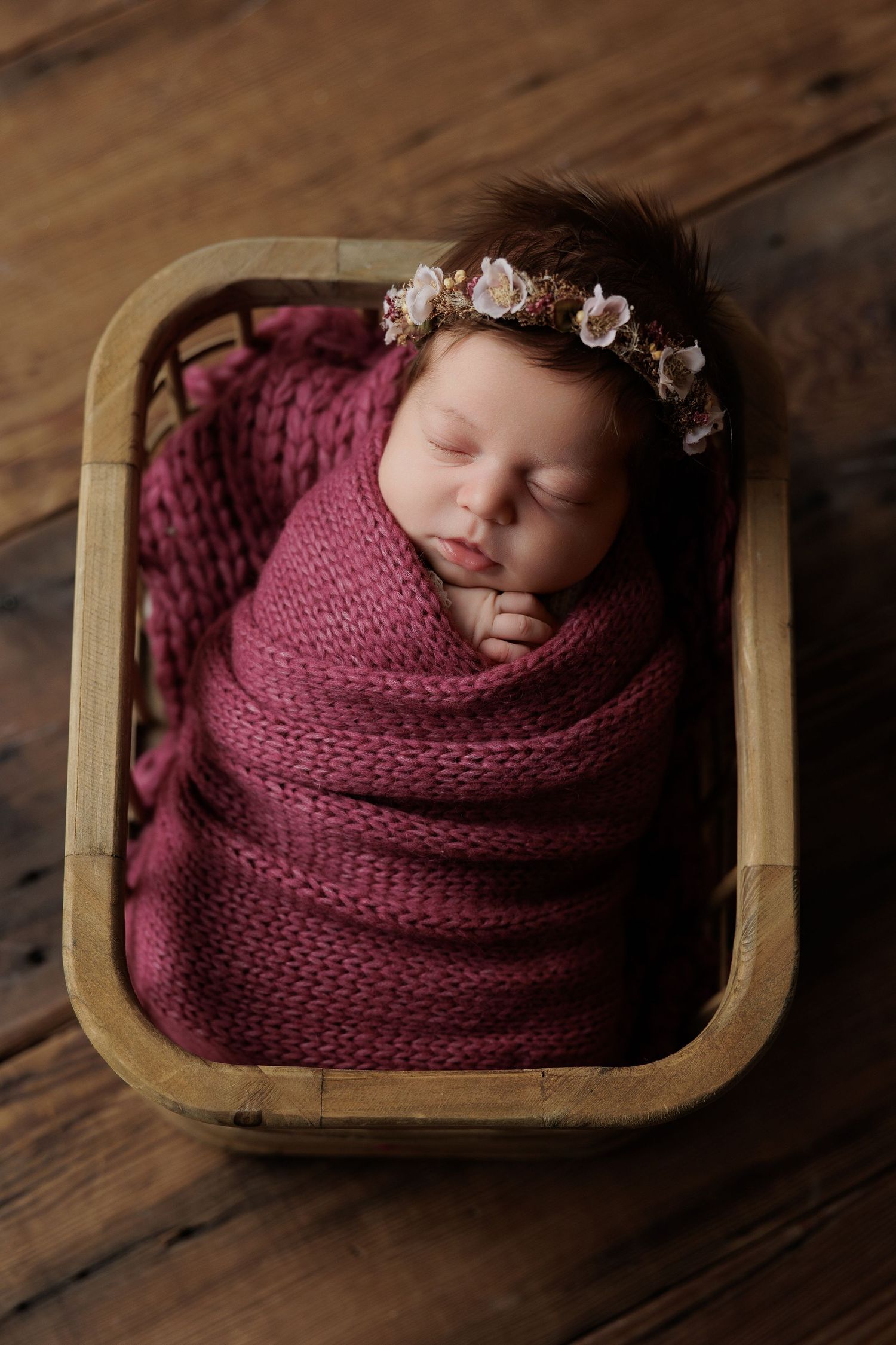 Sleeping newborn swaddled in burgundy knit wrap wearing delicate floral headband rests in wooden basket on hardwood floor.