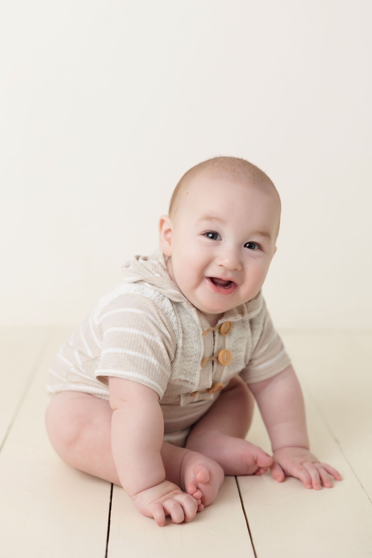 Happy baby wearing beige striped onesie sits on wooden floor with bright expression.