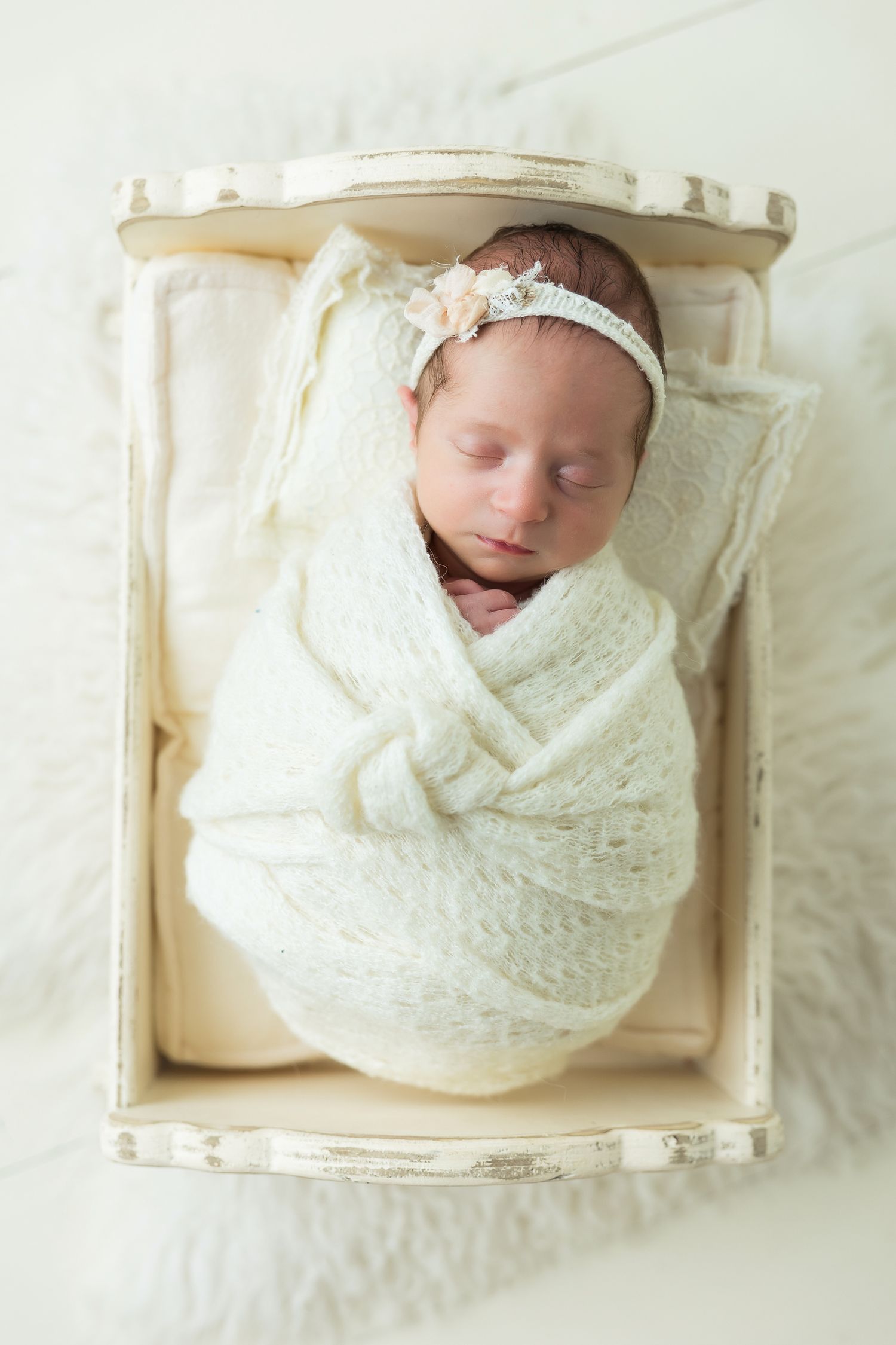 Newborn wrapped in soft white lace swaddle sleeping peacefully in vintage wooden crate with delicate headband.