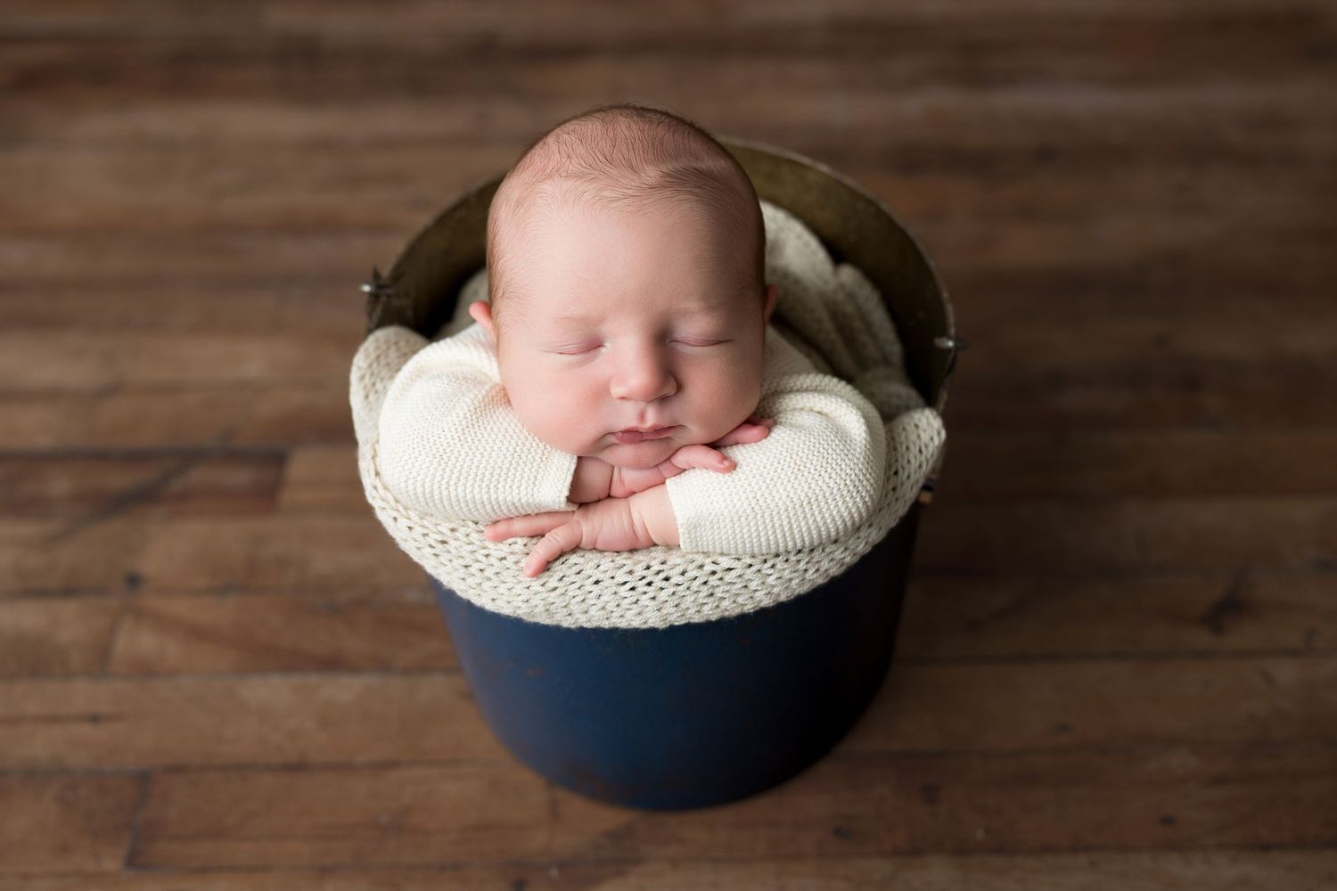 Sleeping newborn rests peacefully in a navy blue bowl lined with soft white fabric on wooden floor.