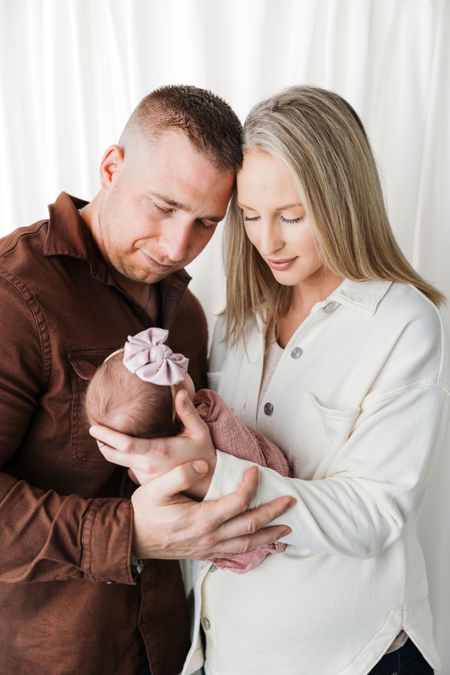 A couple in brown and white sweaters gaze lovingly at their newborn baby wrapped in pink against a light background.