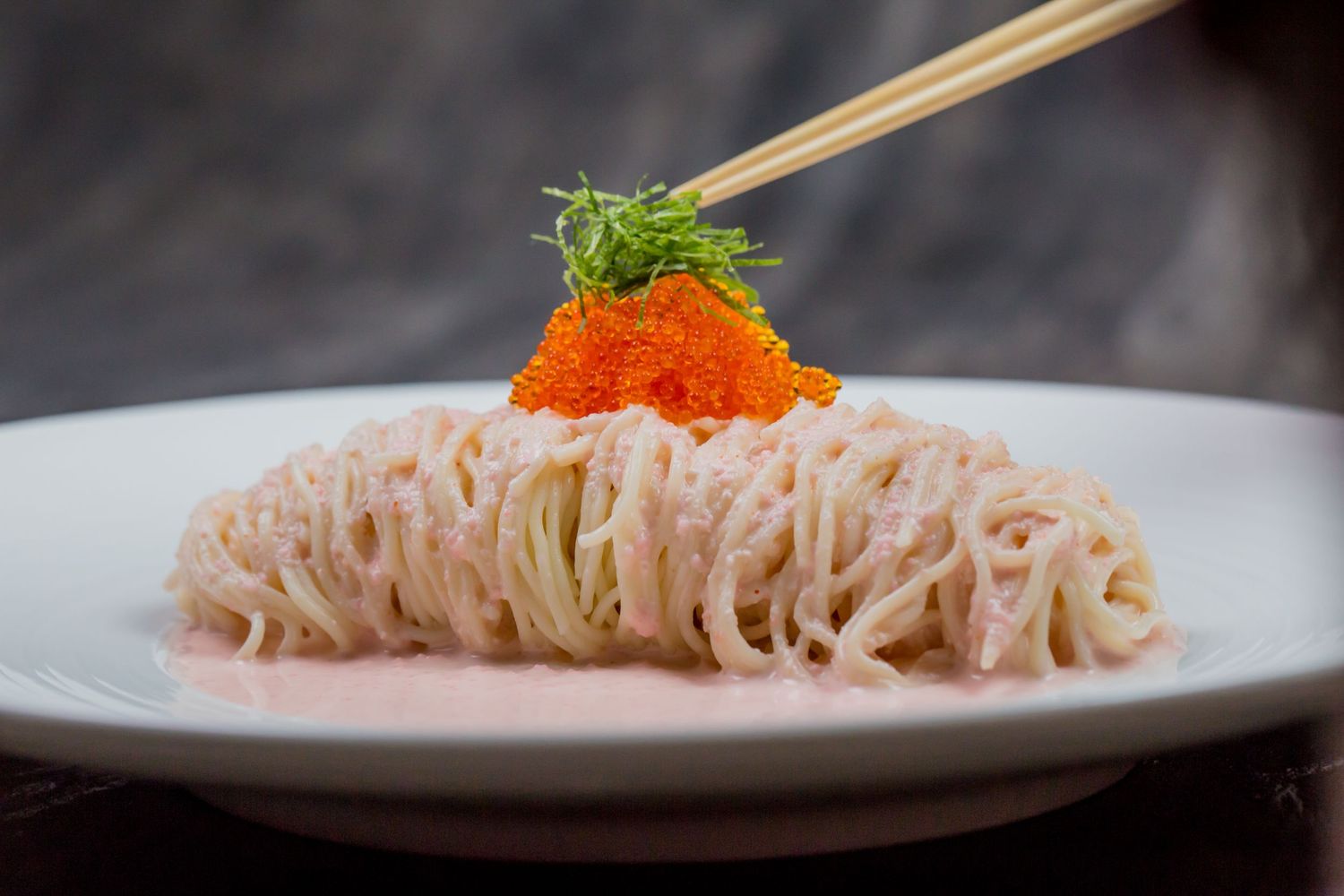 Chopsticks holding fish roe atop creamy pink noodles garnished with green herbs on a white plate.