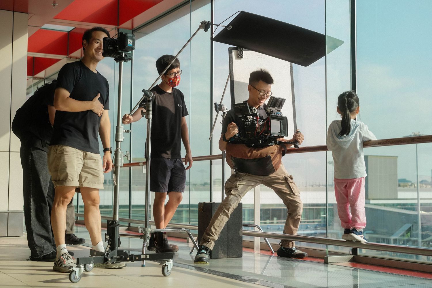 Film crew shooting a guitarist performing near large glass windows in a modern indoor location.