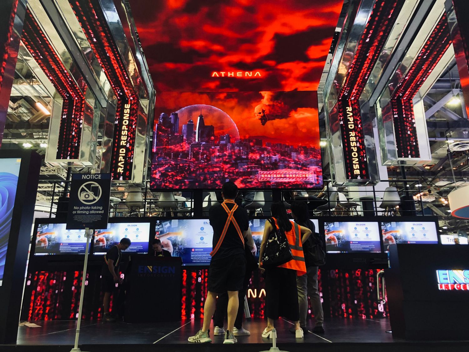 Two visitors explore the Athena gaming exhibit with red-lit displays and dramatic cityscape visuals at a tech expo.