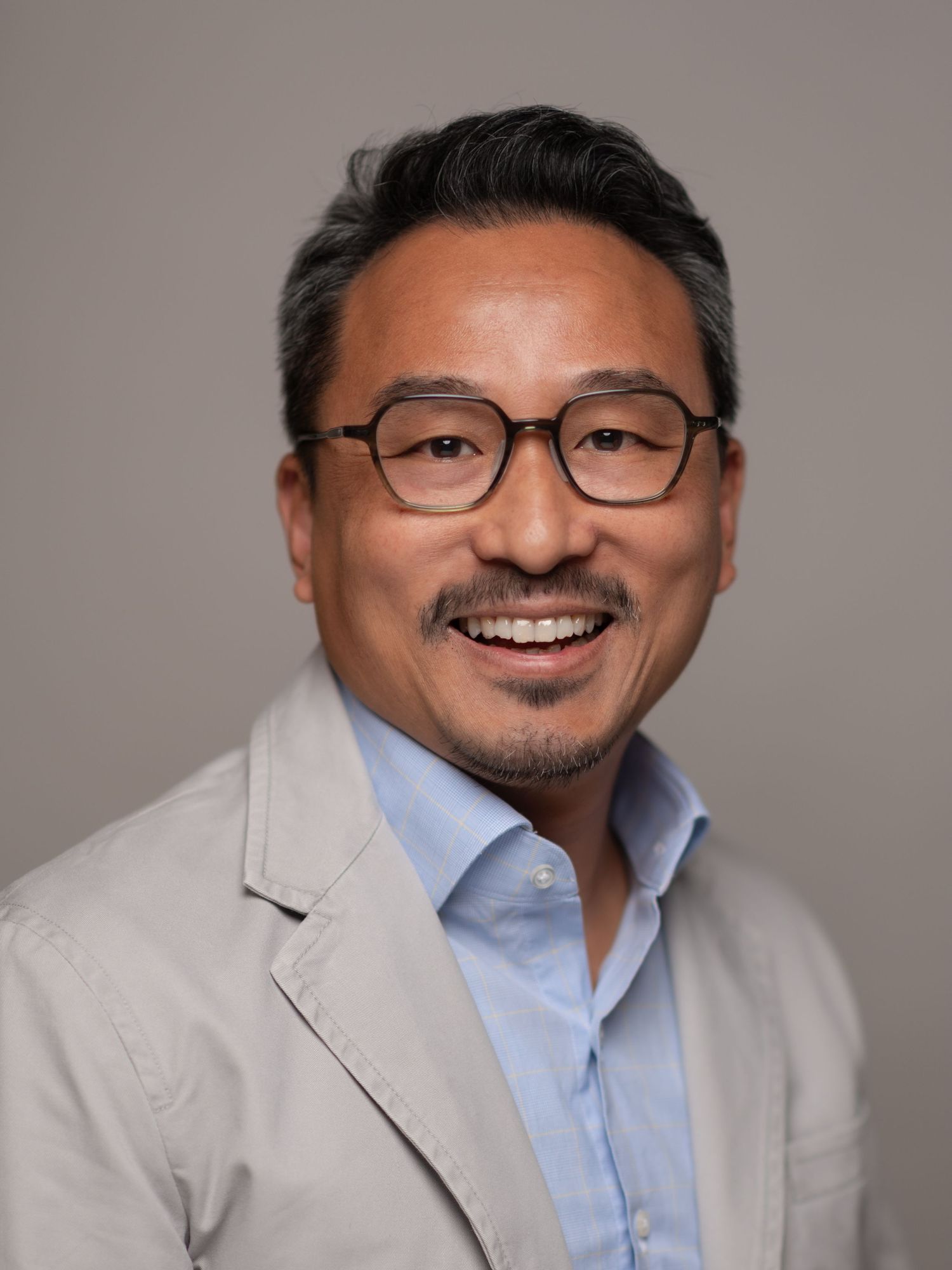 Smiling middle-aged Asian man wearing glasses, light blue shirt, and beige blazer against a gray background.