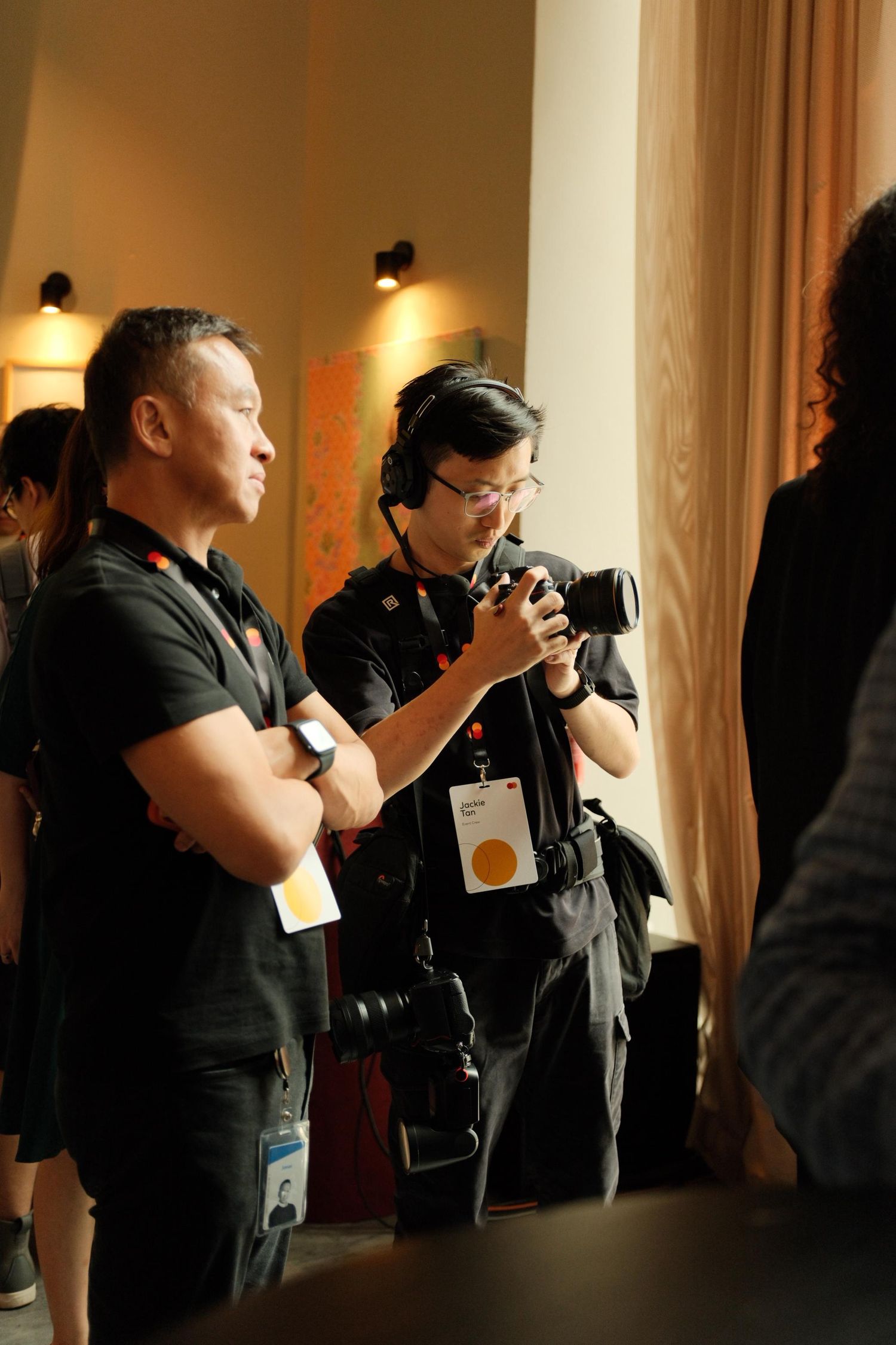 Two photographers in black shirts with lanyards review camera footage at an indoor event venue with warm lighting.