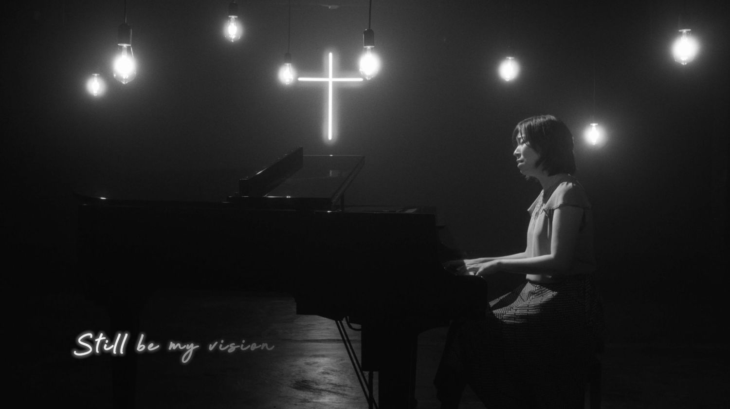 Person playing grand piano in moody black and white setting with cross and hanging lights, text reads Still be my vision.