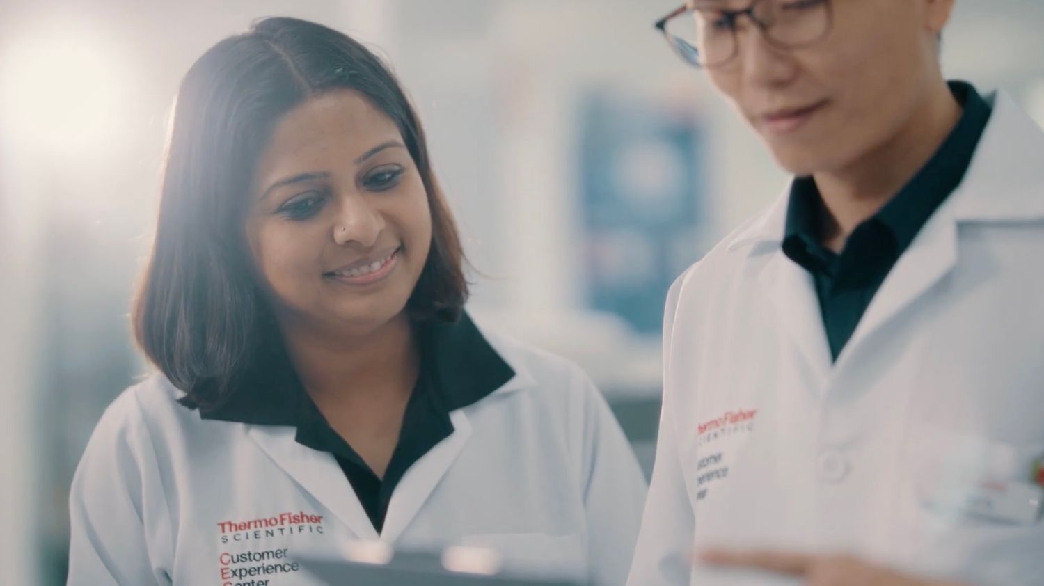 Two female scientists in white lab coats review data together in a bright laboratory setting.