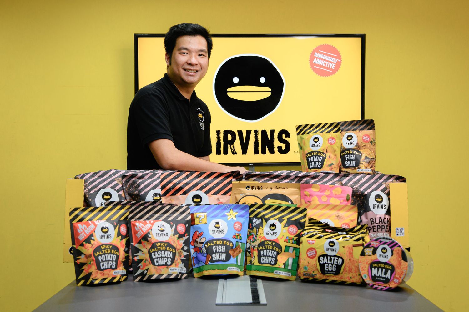 Man posing with IRVINS snack products including salted egg chips displayed on a table against yellow background.