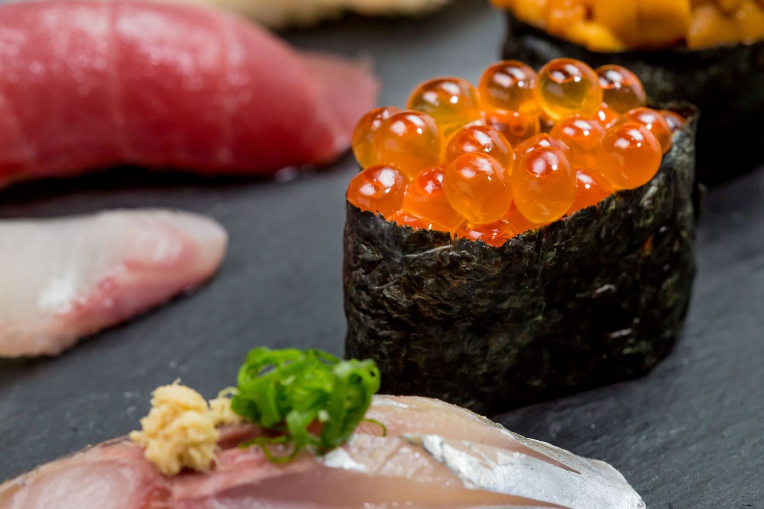 Assorted nigiri and gunkan sushi with salmon roe, tuna, and sea urchin served on a dark slate plate.