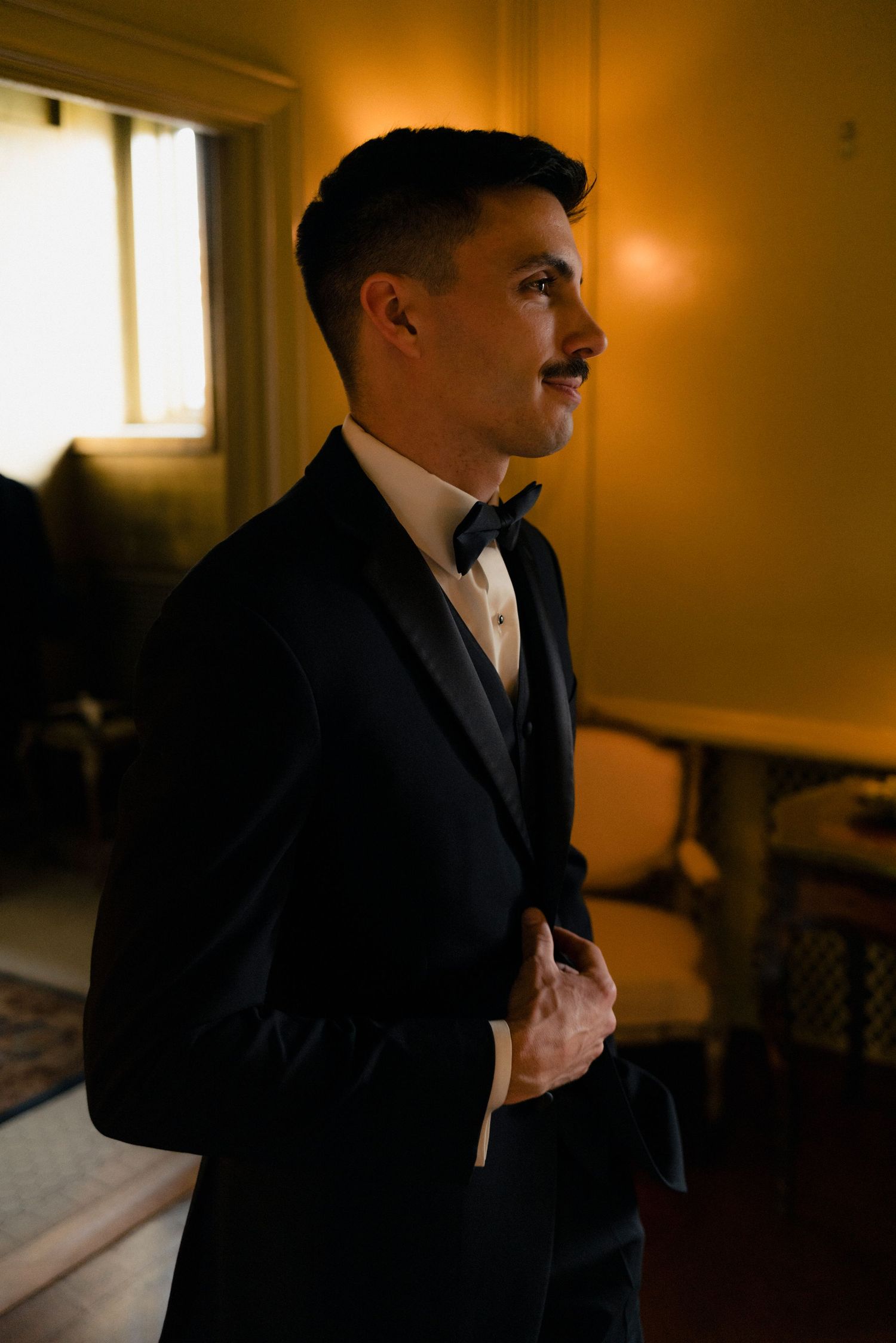 Groom in black tuxedo and bow tie adjusting jacket in warmly lit room before wedding.