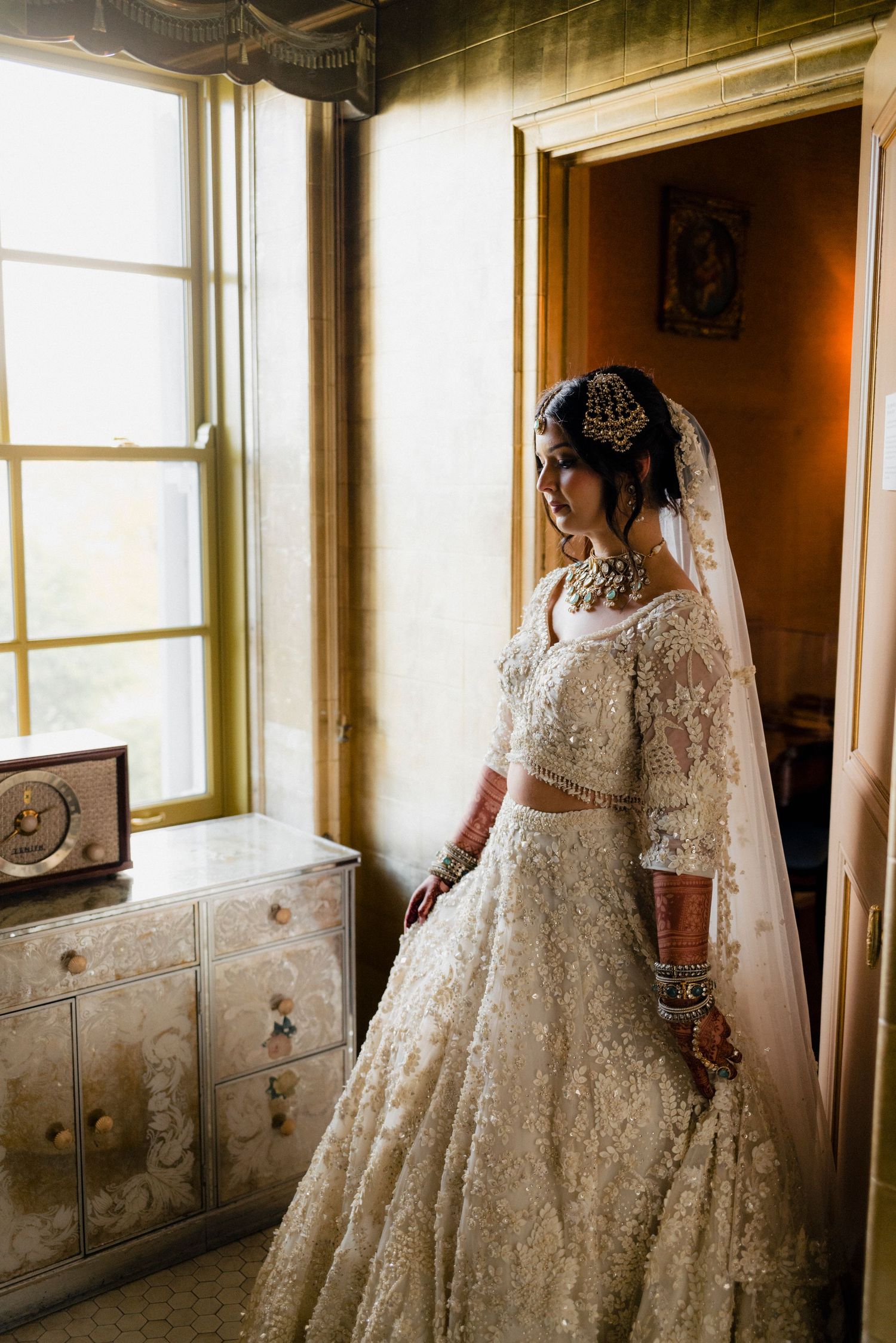 Indian bride in ornate white lehenga and veil standing by window in golden light.