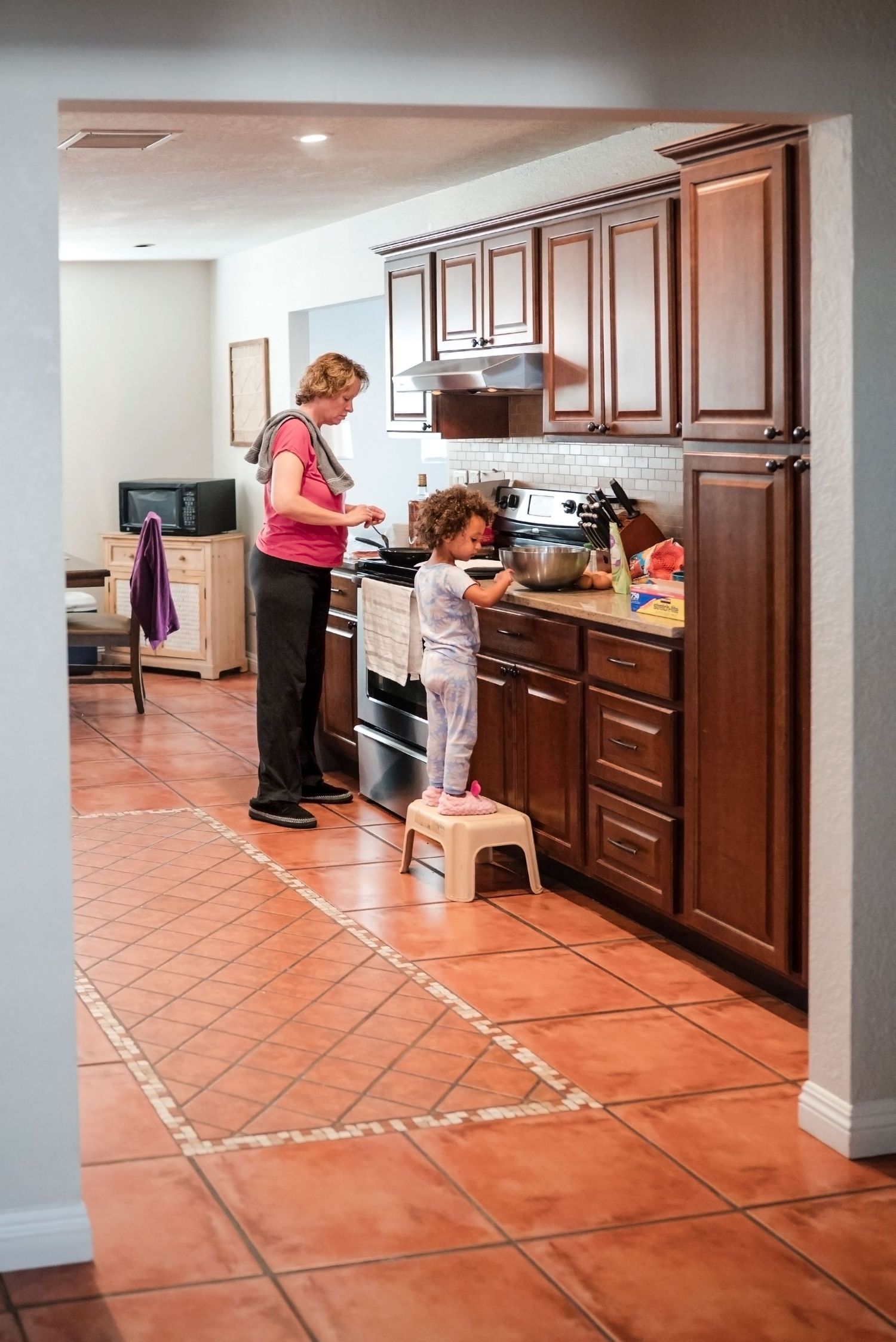 Mother and daughter in kitchen cooking breakfast. Toddler stands on step stool to reach counter. Doughman Photography.