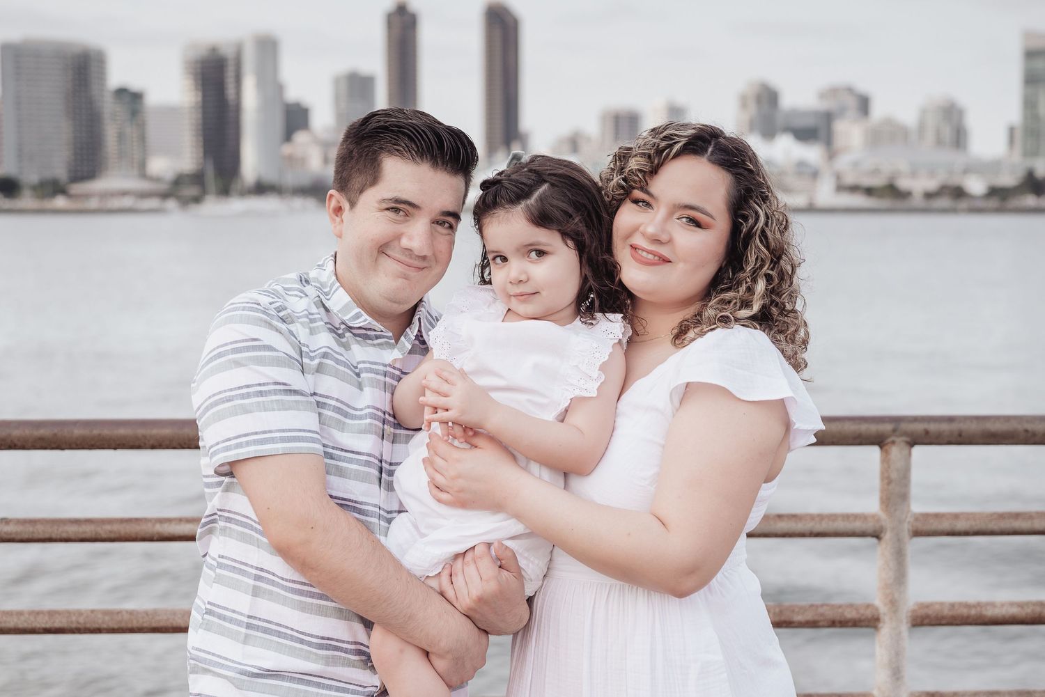 Family standing against boardwalk raining with views of downtown San Diego behind them. Doughman Photography.