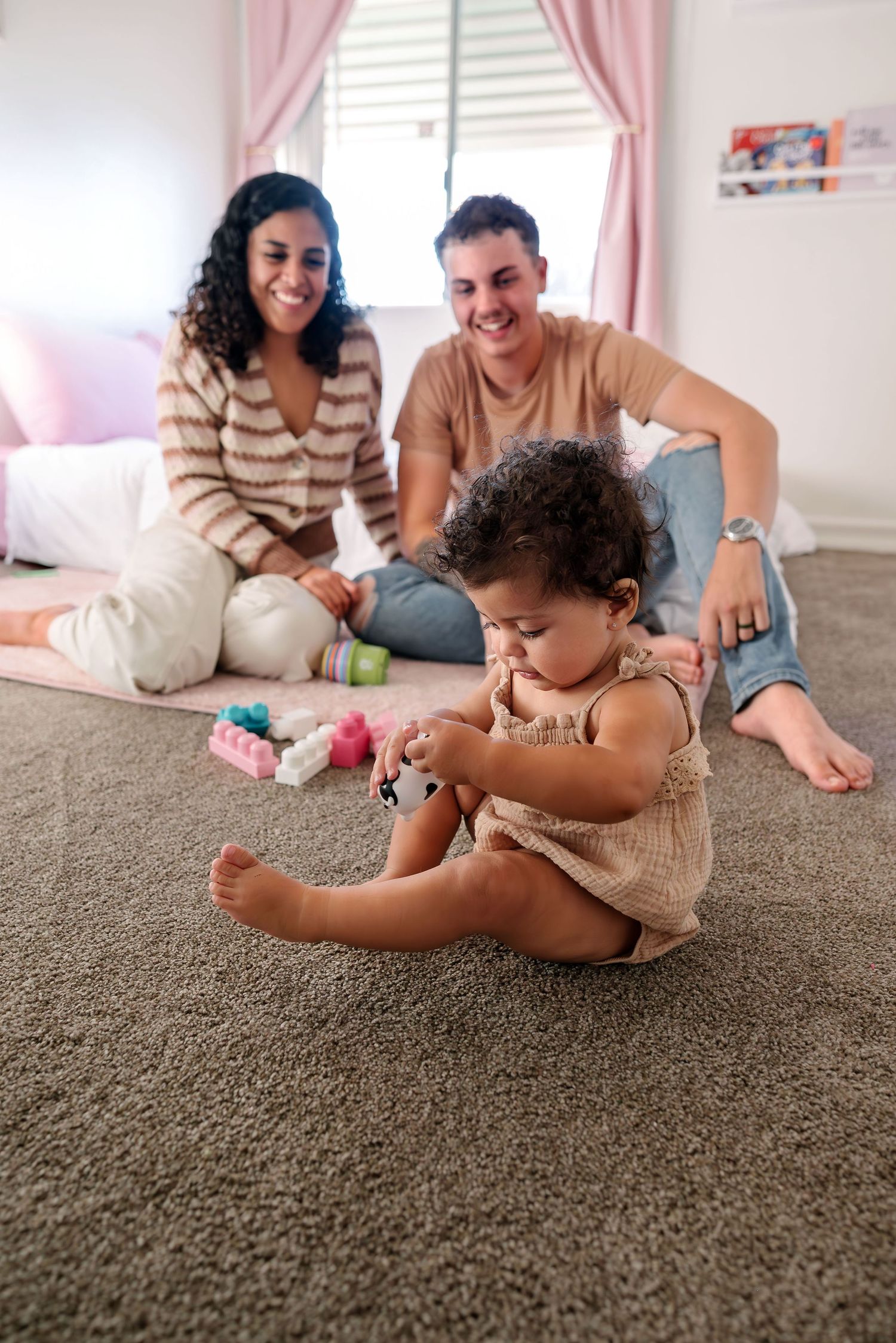 Toddler in focus in the foreground playing with a plastic cow. Parents out of focus in the background smiling at her as she plays. Doughman
