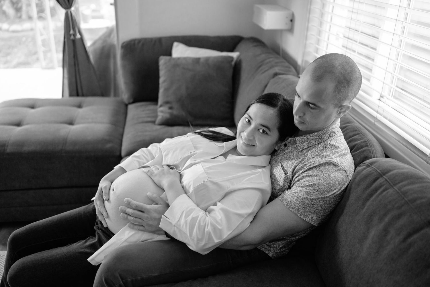 Pregnant woman leaning against her husband as they snuggle on the couch. She looks directly at the camera. He looks at her. black and white