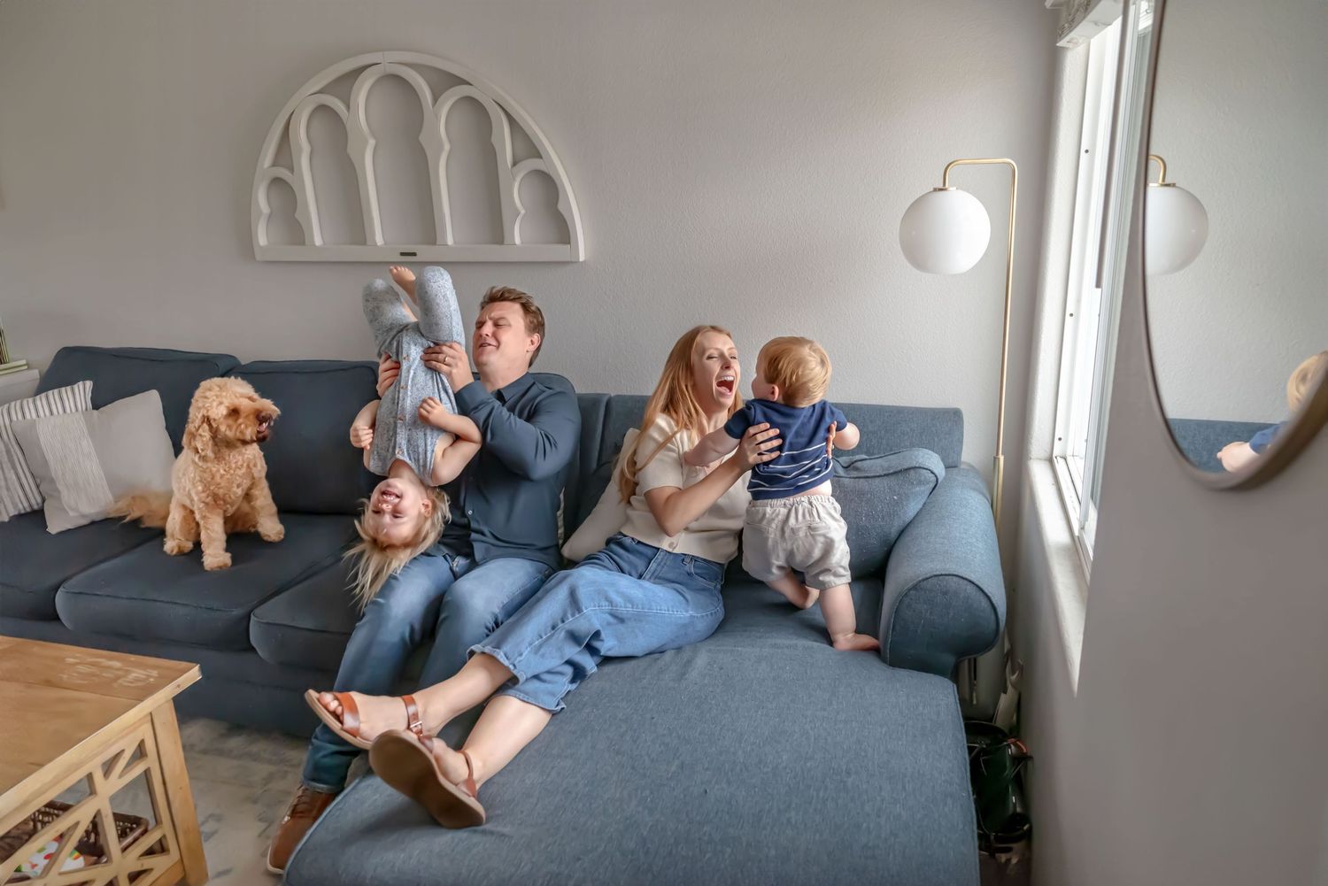 Family sitting on couch. Dad holds toddler upside down. Mom coos at infant. Dog watches. Doughman Photography