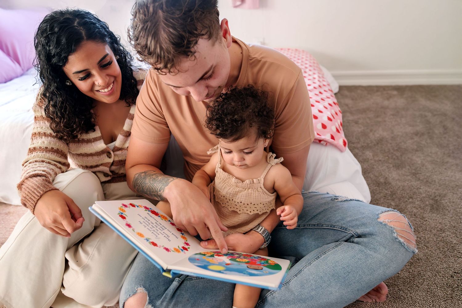 Toddler sitting on dad's lap reading a book. Dad points at the picture. Mom is sitting close by following along, smiling. In-Home Family