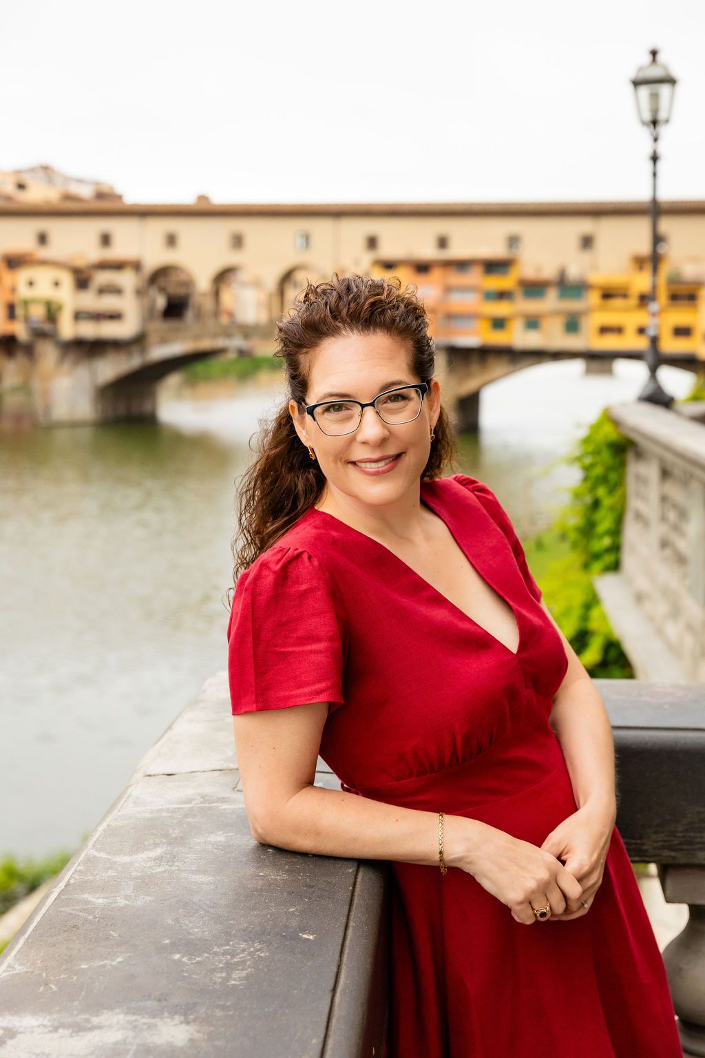 Smiling portrait of Karen Doughman leaning against a low wall with a Venice bridge in the background.  Doughman Photography.