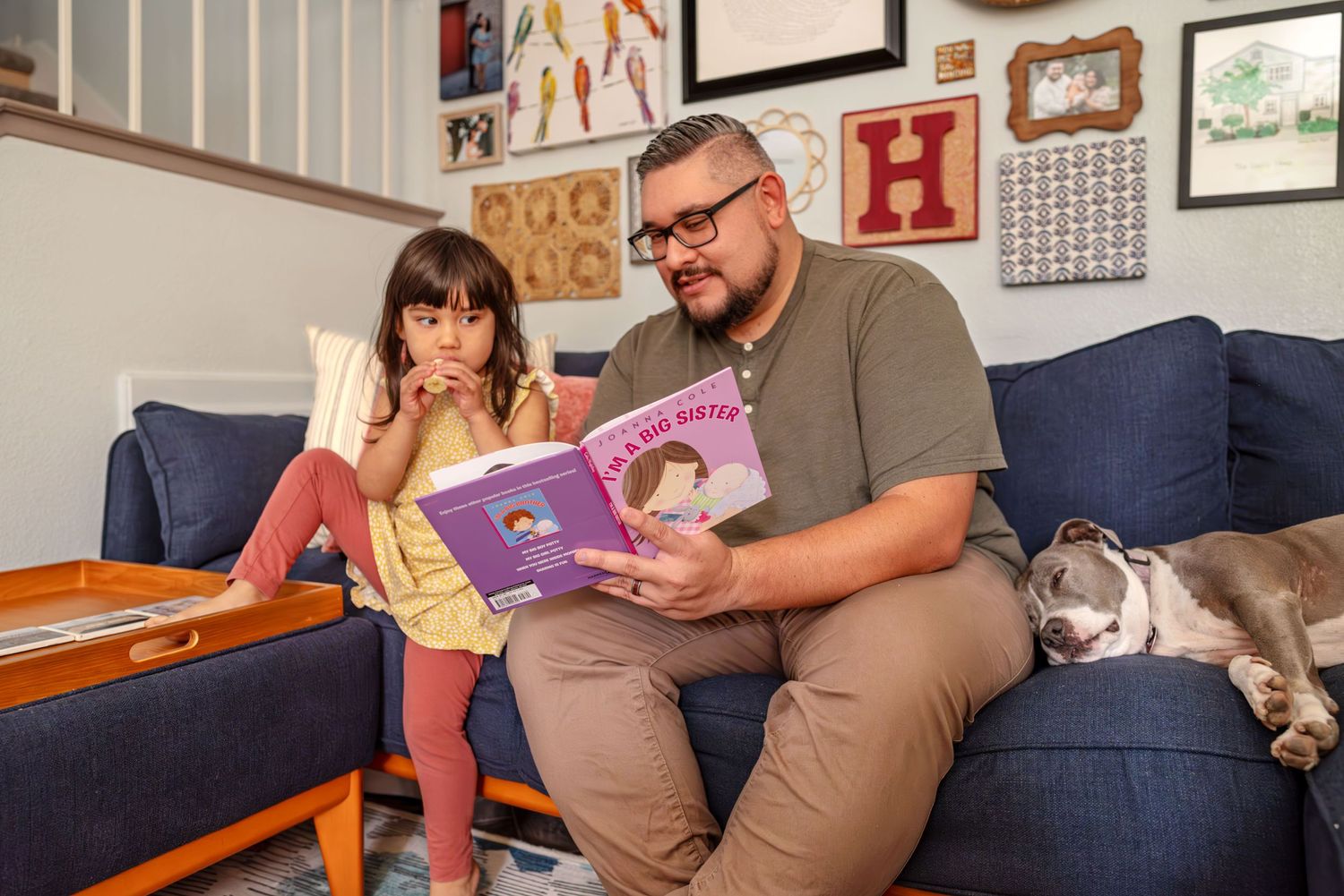 Dad and toddler sit on couch and reads to his toddler a book about being a big sister. Toddler eats a snack. Dog slumbers next to them.