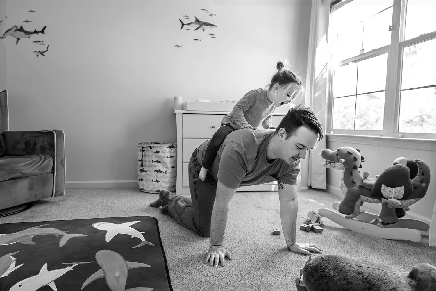Father giving his daughter a piggyback ride in a bright nursery, surrounded by shark-themed decor and toys. Doughman Photography