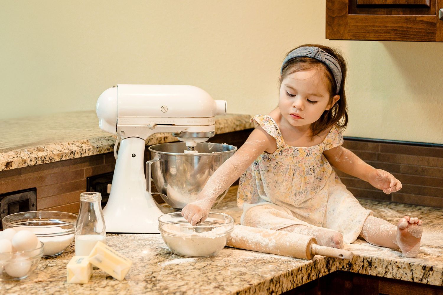 Toddler sitting on kitchen counter next to mixer. Flour is everywhere - counter, her face, her dress. In-Home Session Activity. Doughman