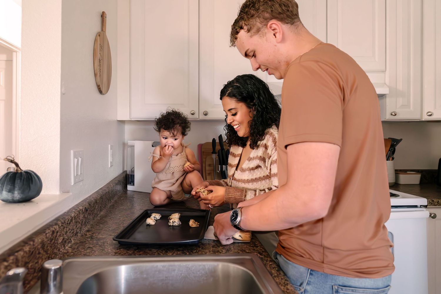 Parents placing cookie dough on baking tray while young toddler steals one. 