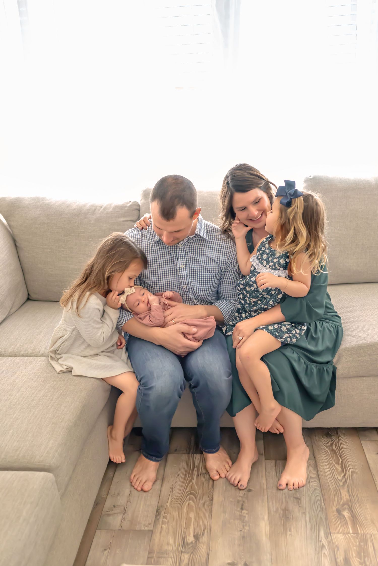 A family of five cuddles on the couch, with two sisters adoring their newborn sibling as parents share loving smiles. Doughman Photography