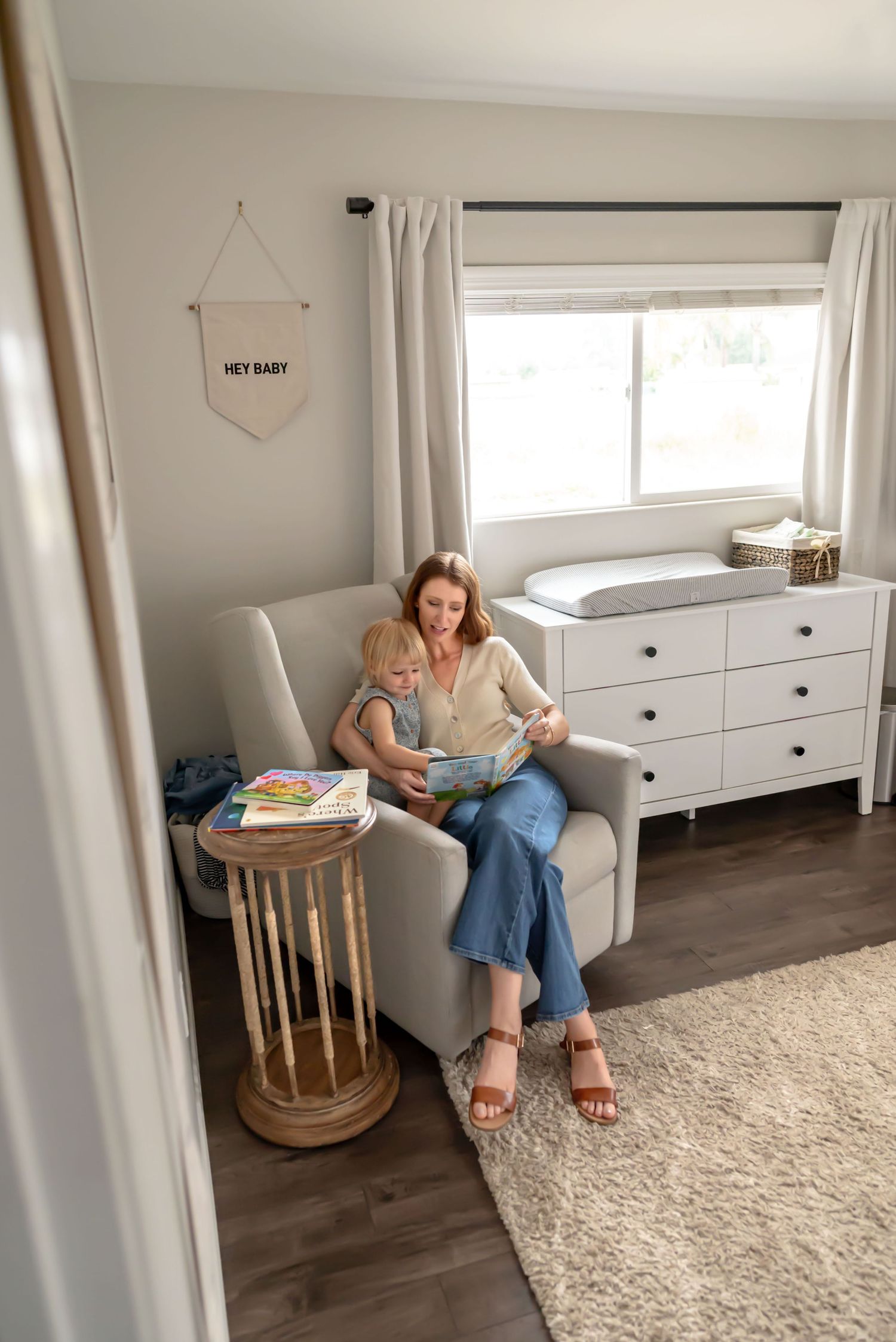 Mother and toddler cuddle in a cozy nursery, reading a book together in a soft armchair near a window. Doughman Photography
