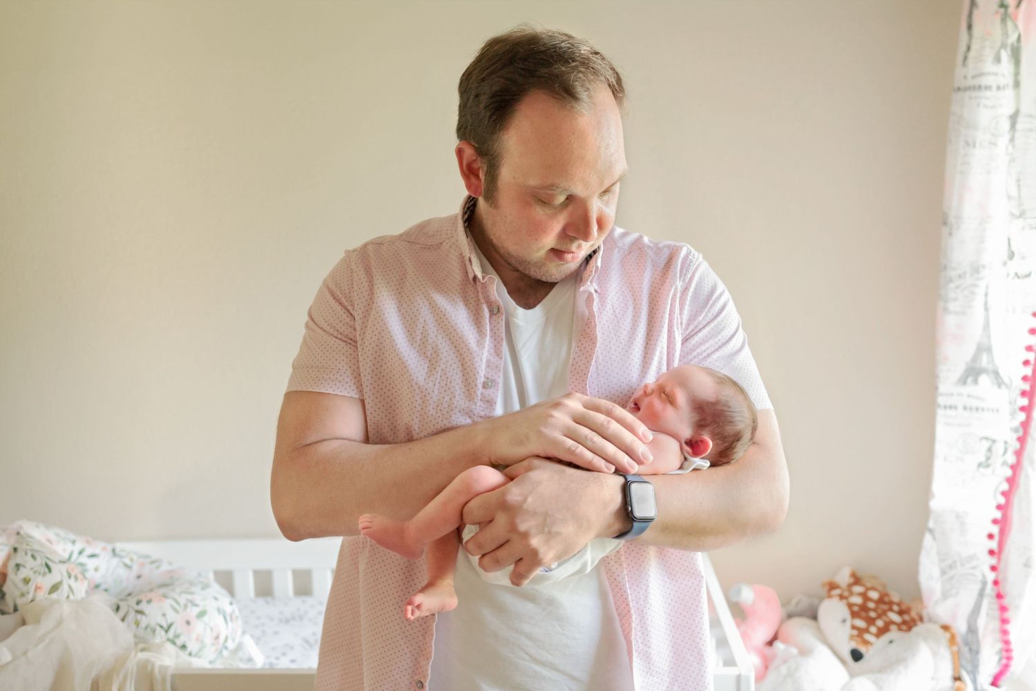 Dad standing in nursery looking down at his newborn daughter cradled in his arms. Newborn In-Home Family Session. Doughman Photography.